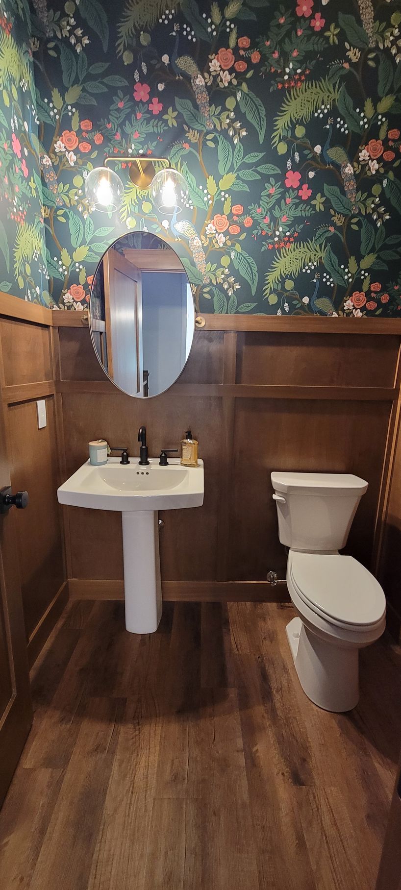 Powder room with floral wallpaper, wooden paneling, pedestal sink, and toilet.