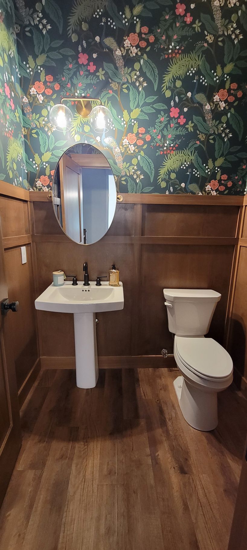 A small bathroom with floral wallpaper, a pedestal sink, and a toilet. The walls are wood-paneled.