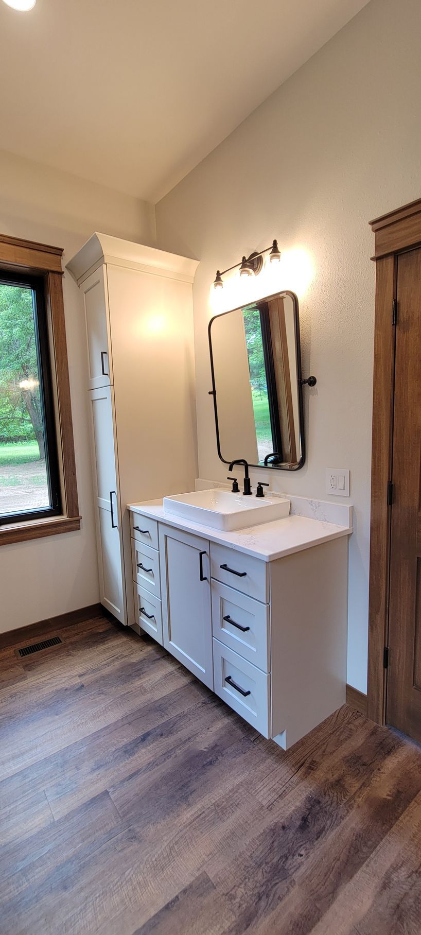 Bathroom with white vanity, mirror, and tall cabinet against a light-colored wall; wood-look flooring.