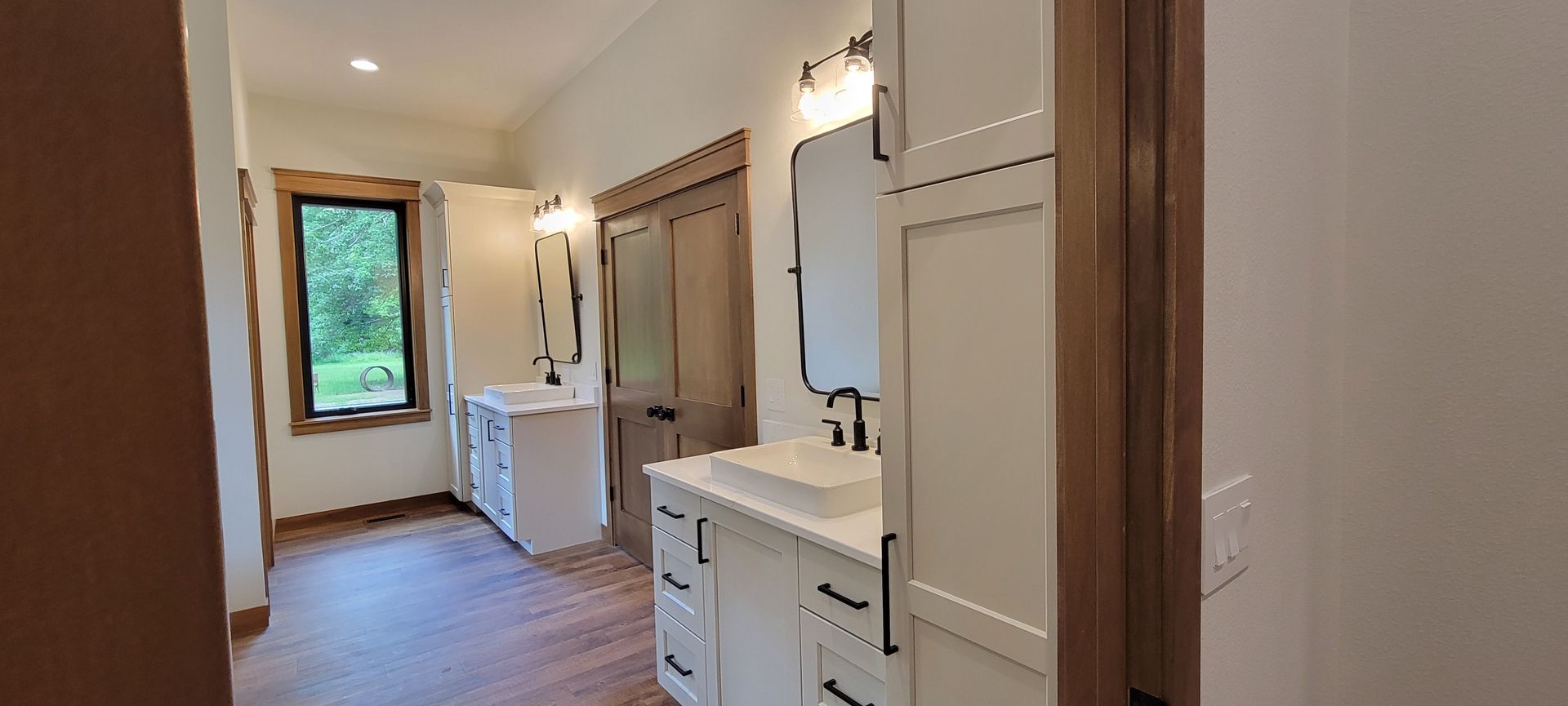 Bathroom with white cabinets, dark hardware, wood trim, and a window overlooking greenery.