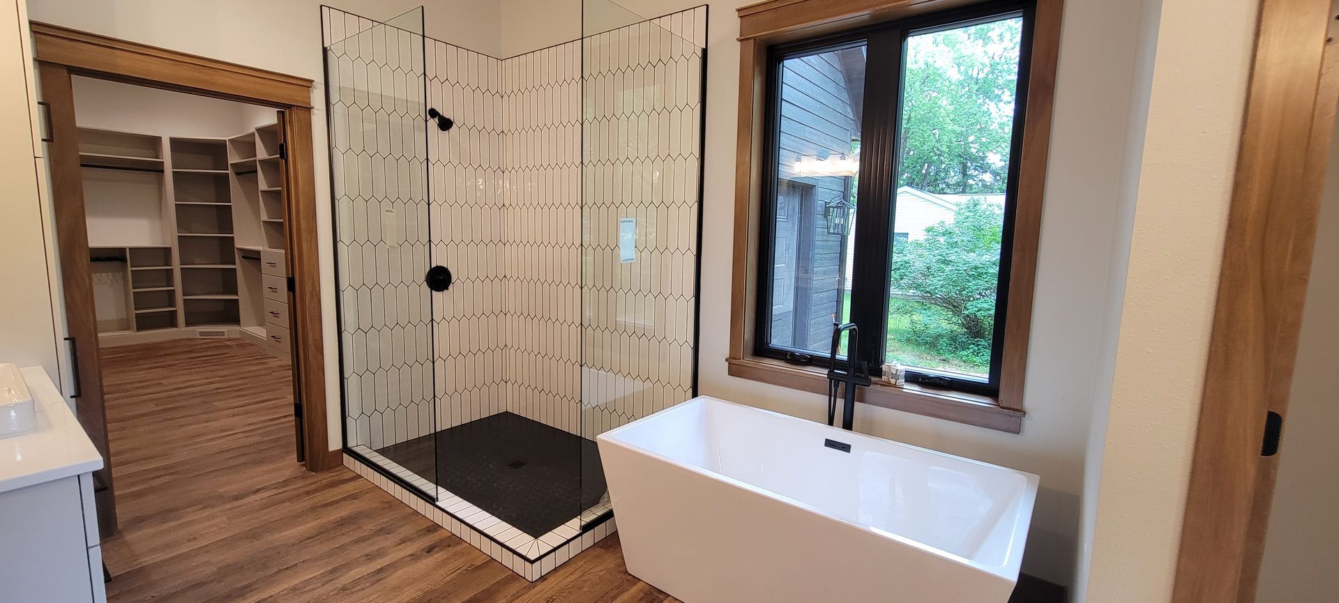 Bathroom with shower, tub, walk-in closet, and a window overlooking greenery. Wooden accents and neutral tones.