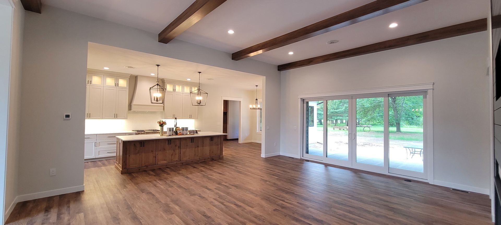 Open-concept interior with kitchen island, wood floors, white walls, and exposed wooden beams. Sliding glass doors lead to outdoors.