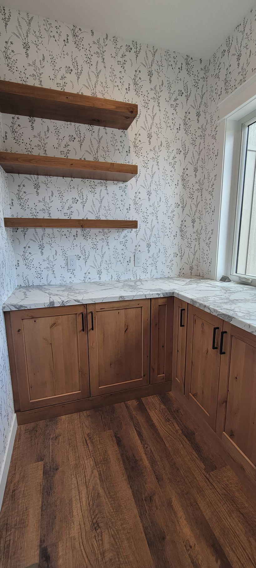 Wooden cabinets, countertops, and open shelving in a kitchen nook with speckled wallpaper and hardwood floors.