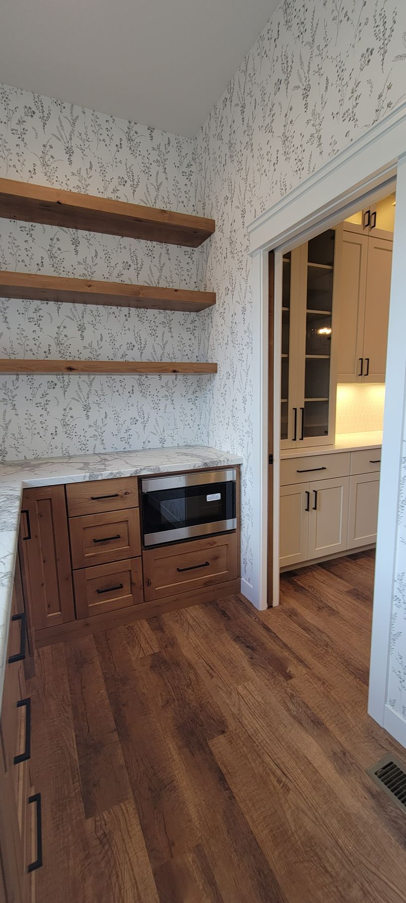 Kitchen pantry with shelves, microwave, wooden cabinets, and a doorway to another kitchen area.