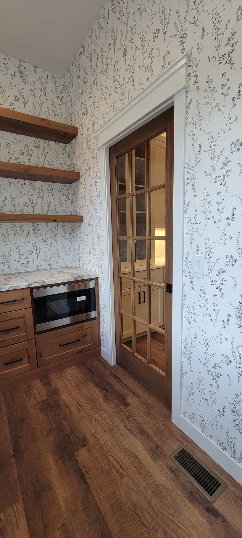 Interior of a kitchen pantry with wood shelving, cabinetry, and flooring. A glass-paned door opens into a bright area.