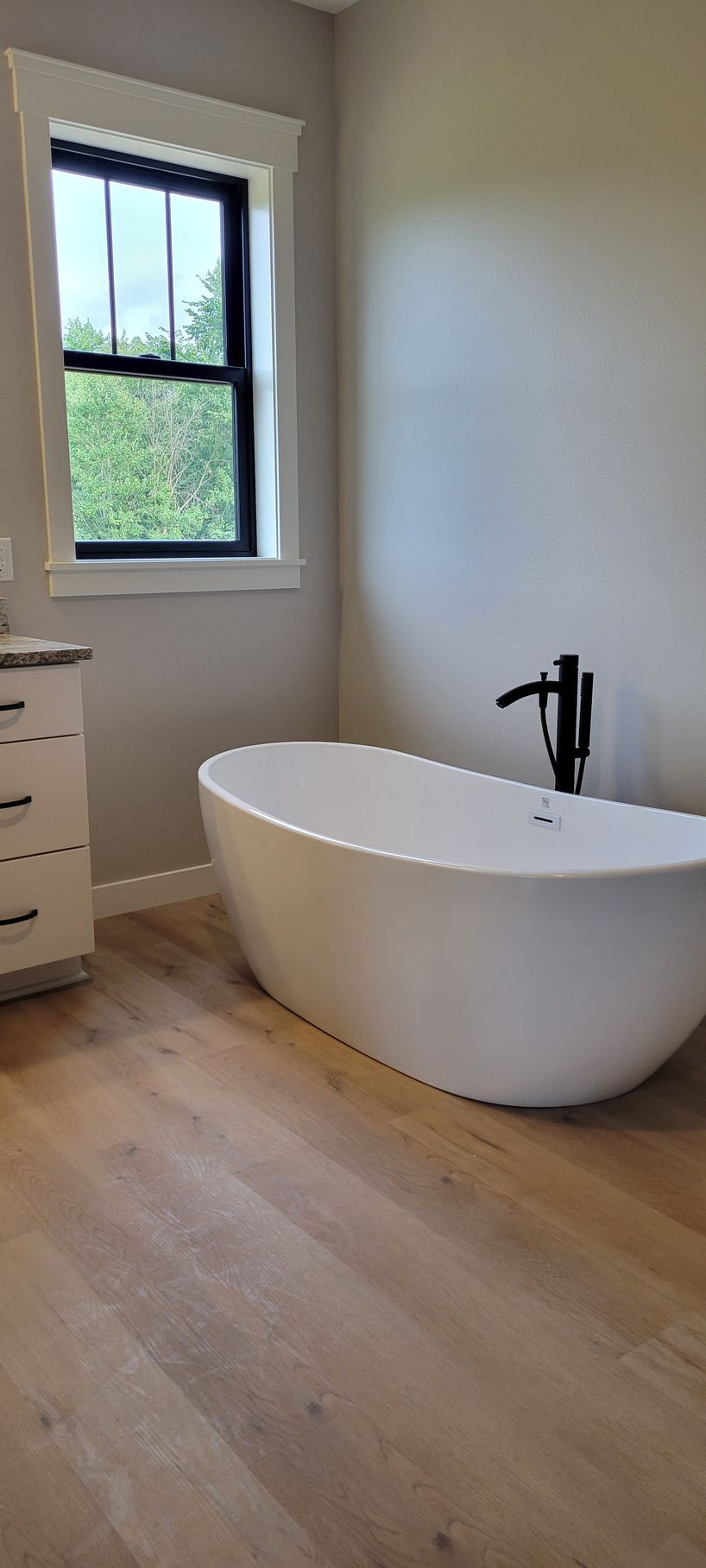 Bathroom with a freestanding oval bathtub, black faucet, and a window with black trim. Light wood-look flooring.
