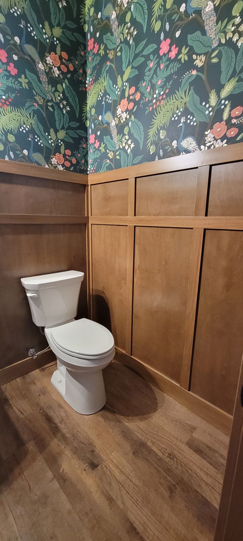 A bathroom with a white toilet, wood paneling, floral wallpaper, and wood-look flooring.