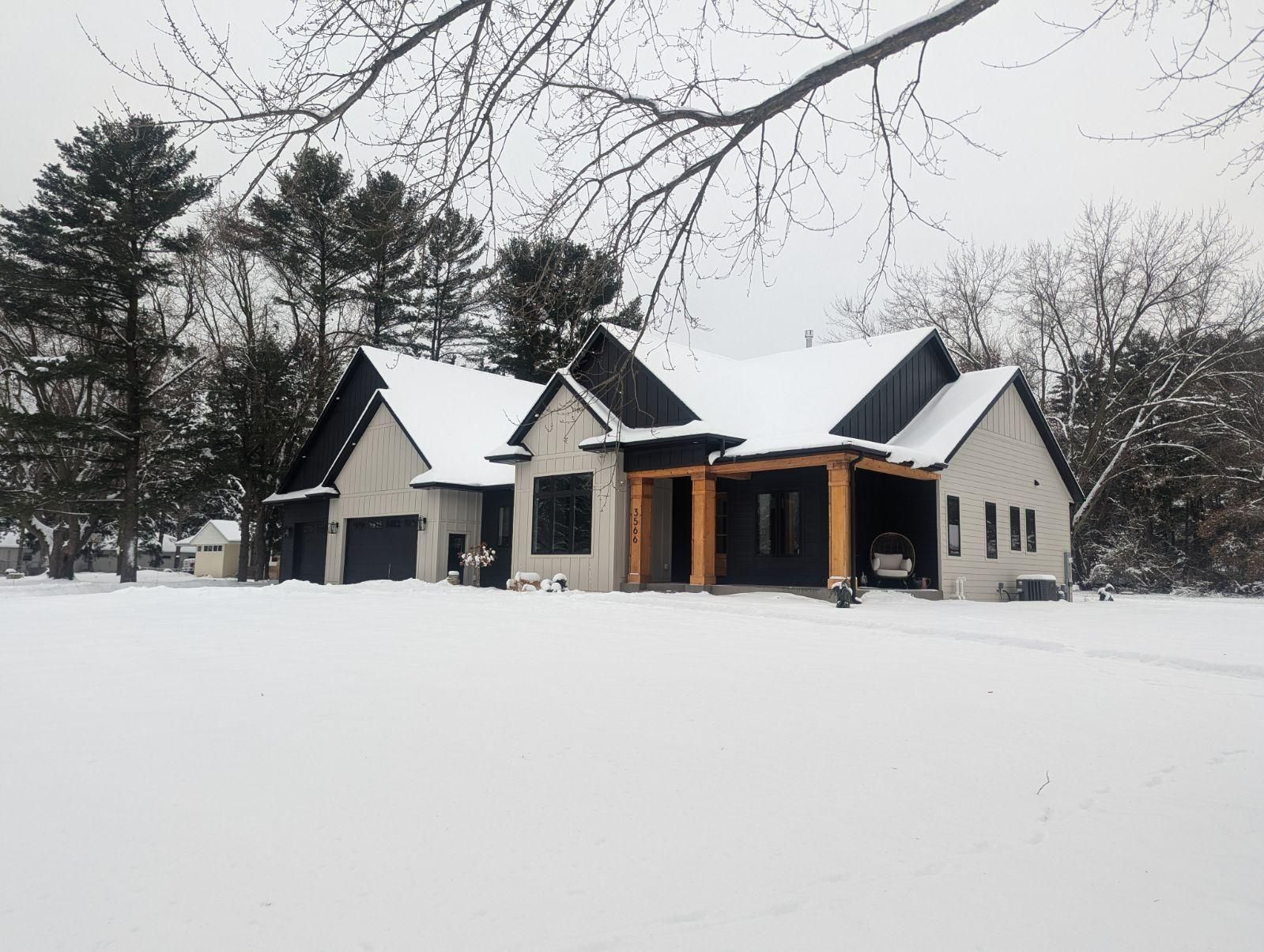 Snow-covered house with black roof and white siding, surrounded by trees under a cloudy sky.