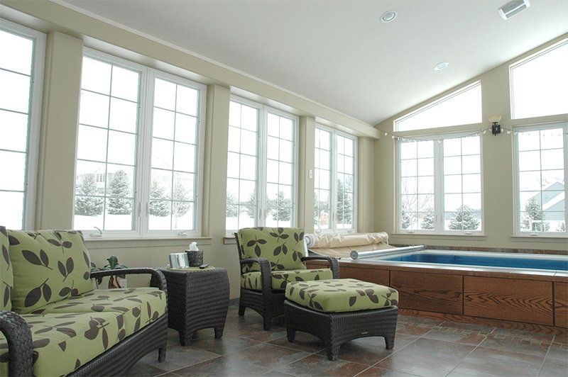 Bright sunroom with green patterned chairs, ottoman, and large windows overlooking a wooden deck