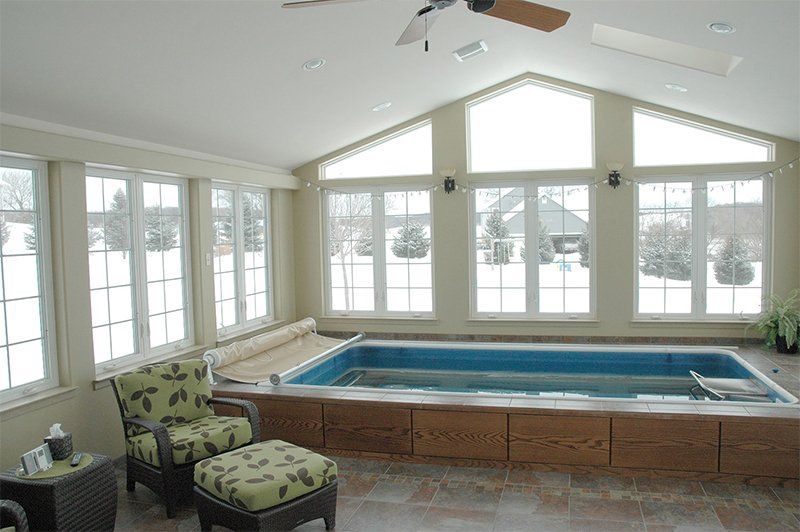 Bright sunroom with large windows, a hot tub, and cushioned chairs overlooking snowy outdoors