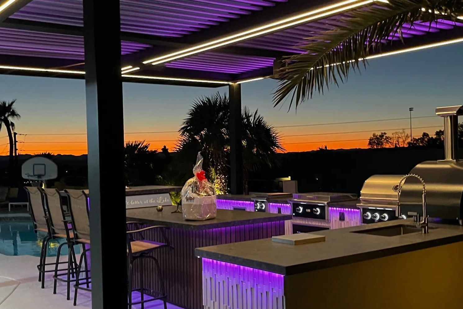 Outdoor bar at sunset with purple-lit pergola, and poolside seating