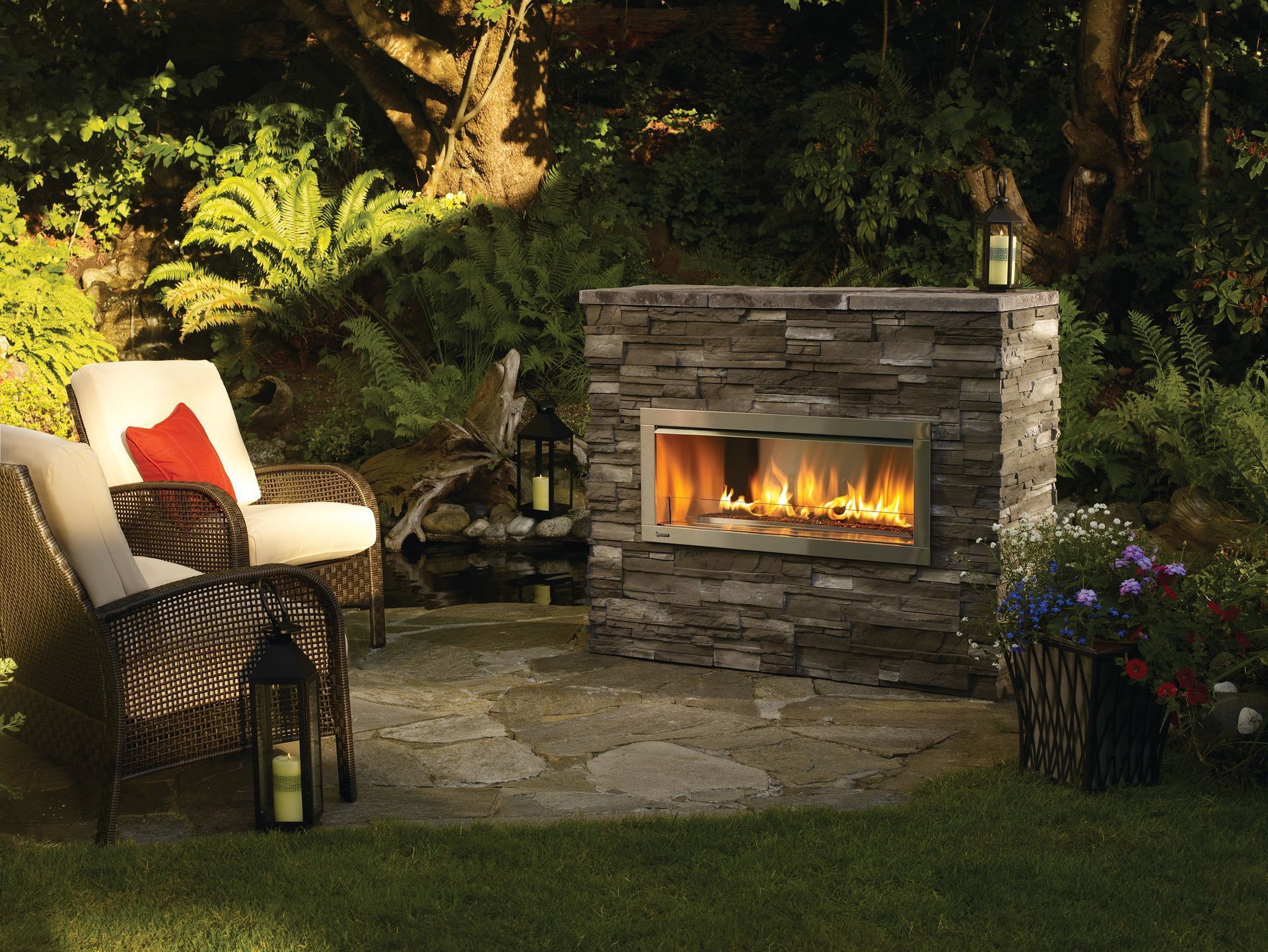 Outdoor stone fireplace with flames beside a cushioned chair and potted flowers in a garden patio
