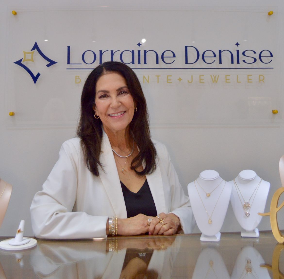 A woman in a white blazer sits at a jewelry store counter, backed by a wall sign reading Lorraine Denise Jewelry.