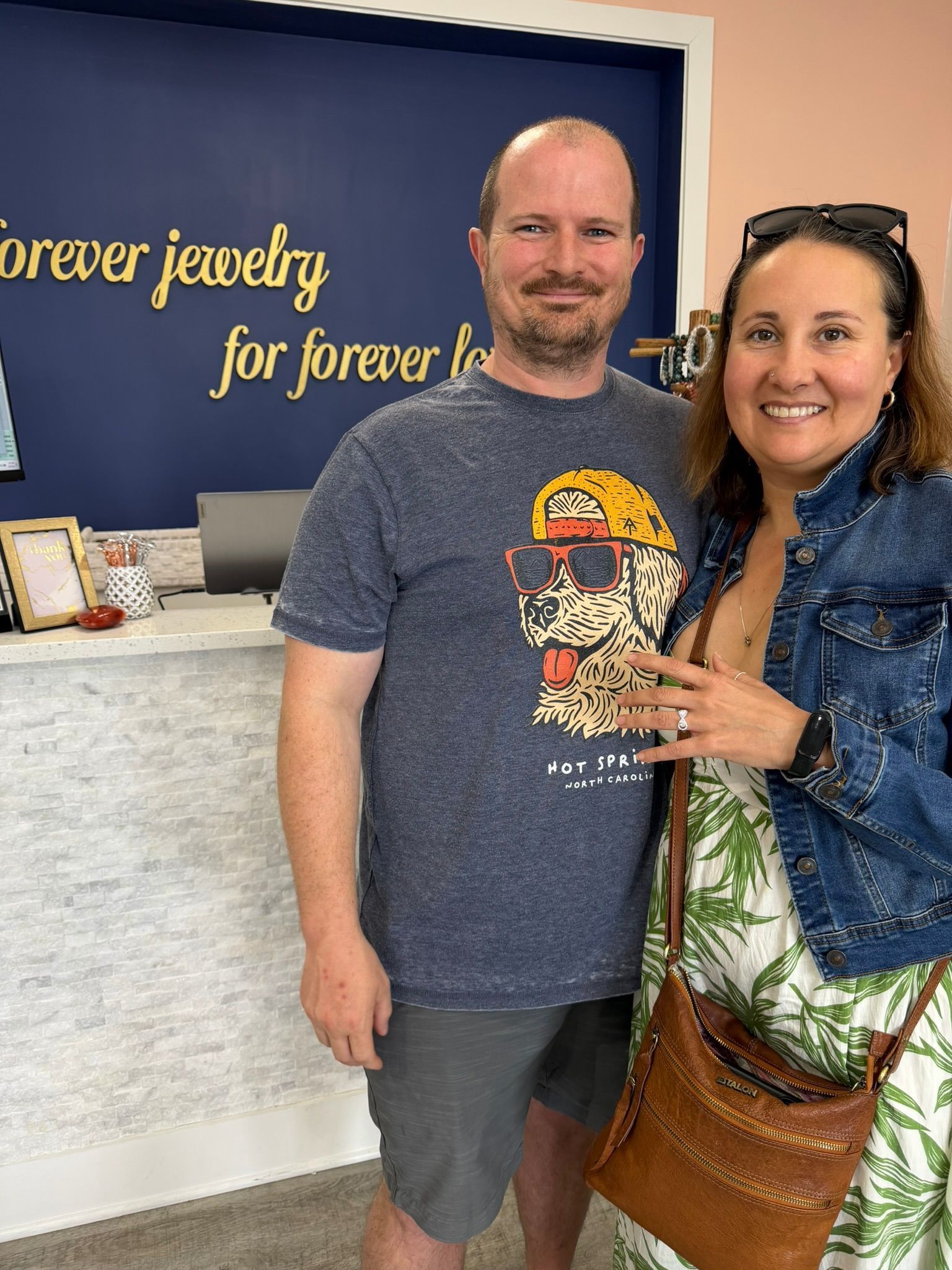 Two smiling people stand in a jewelry shop, one holding a small bouquet of flowers.