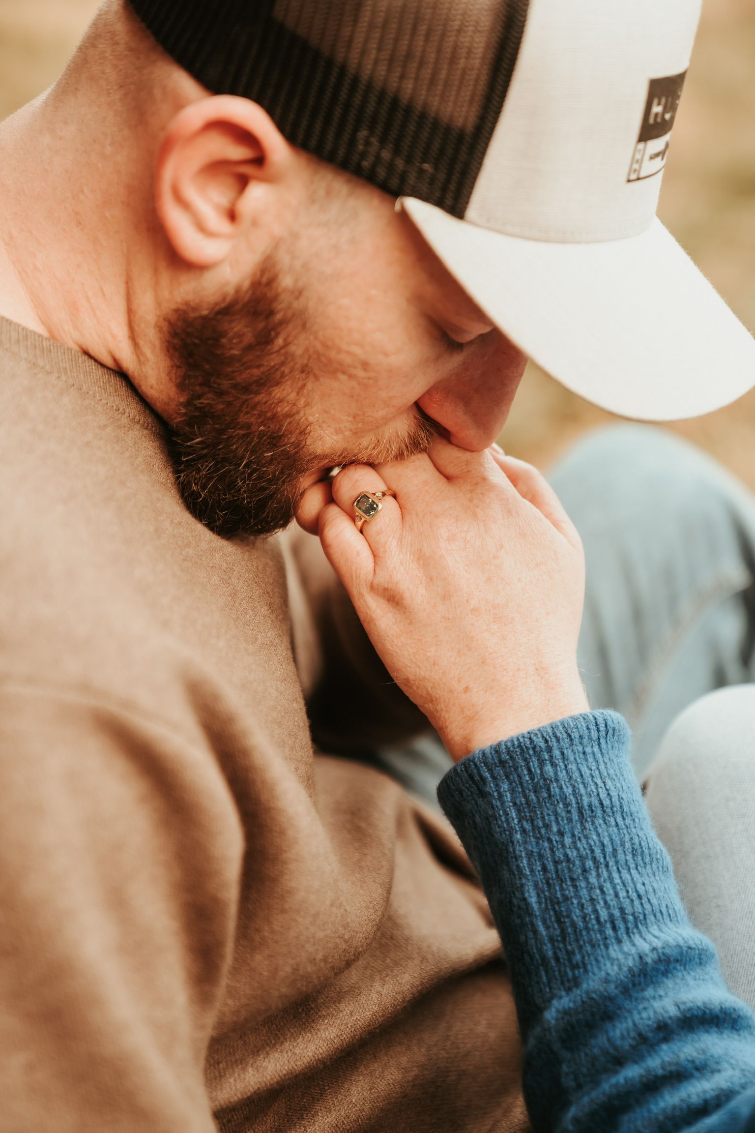 Couple embracing closely, man in tan sweater and white cap, woman’s hand on his face.