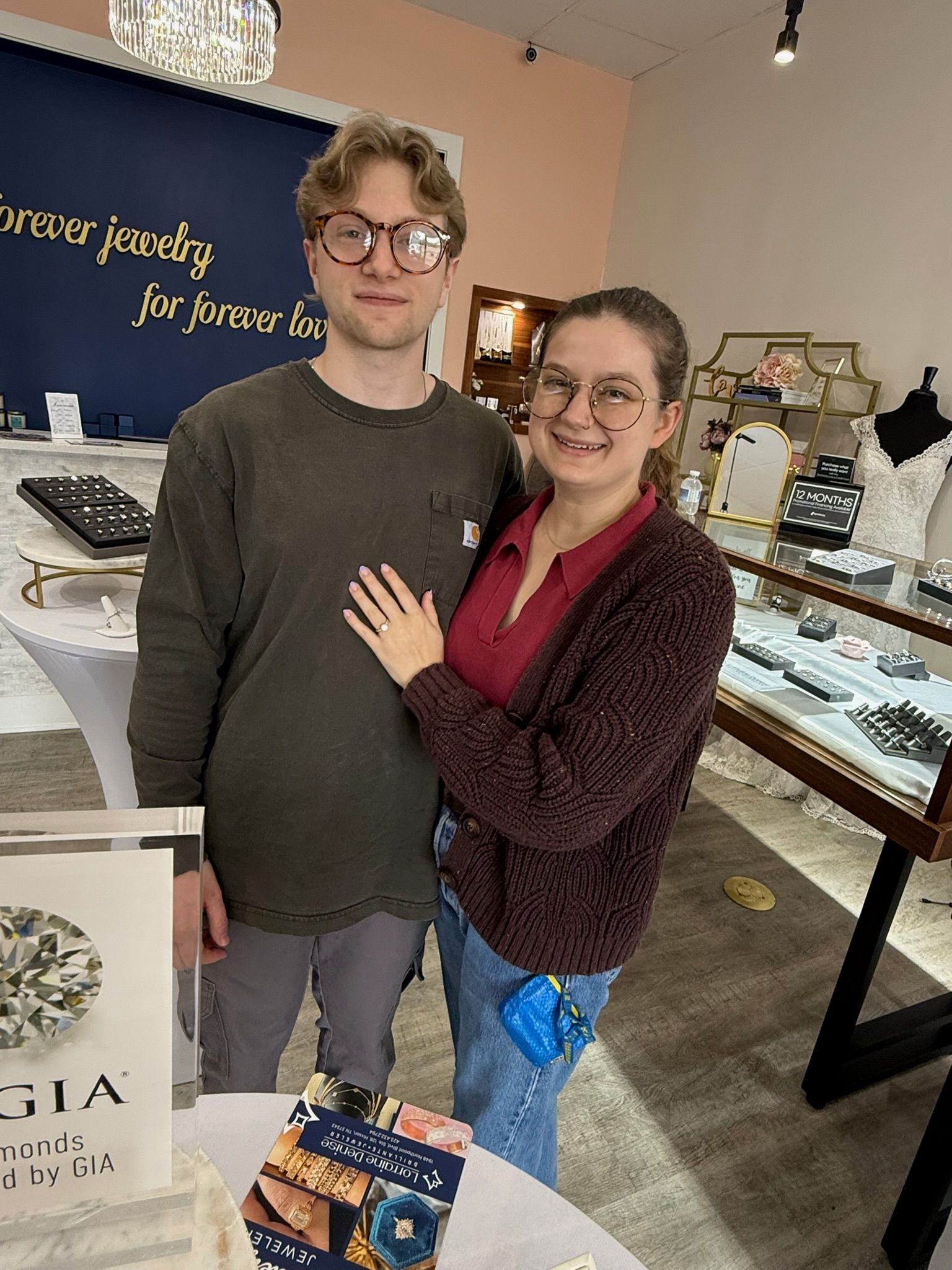 Two people posing in a jewelry store, smiling beside glass display cases.