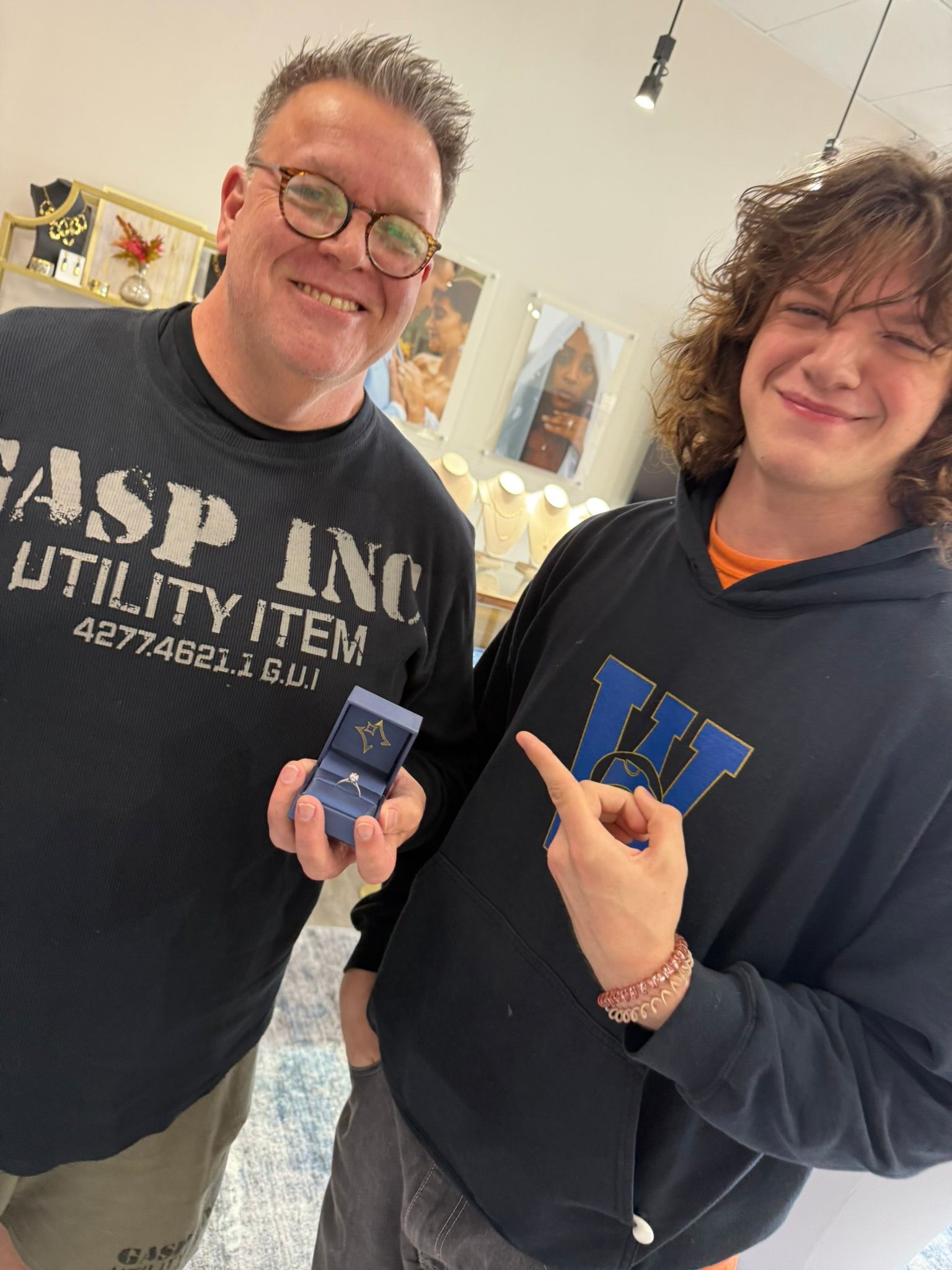 Two smiling people indoors, holding a ring and pointing at it, one in a black shirt and one in a navy hoodie