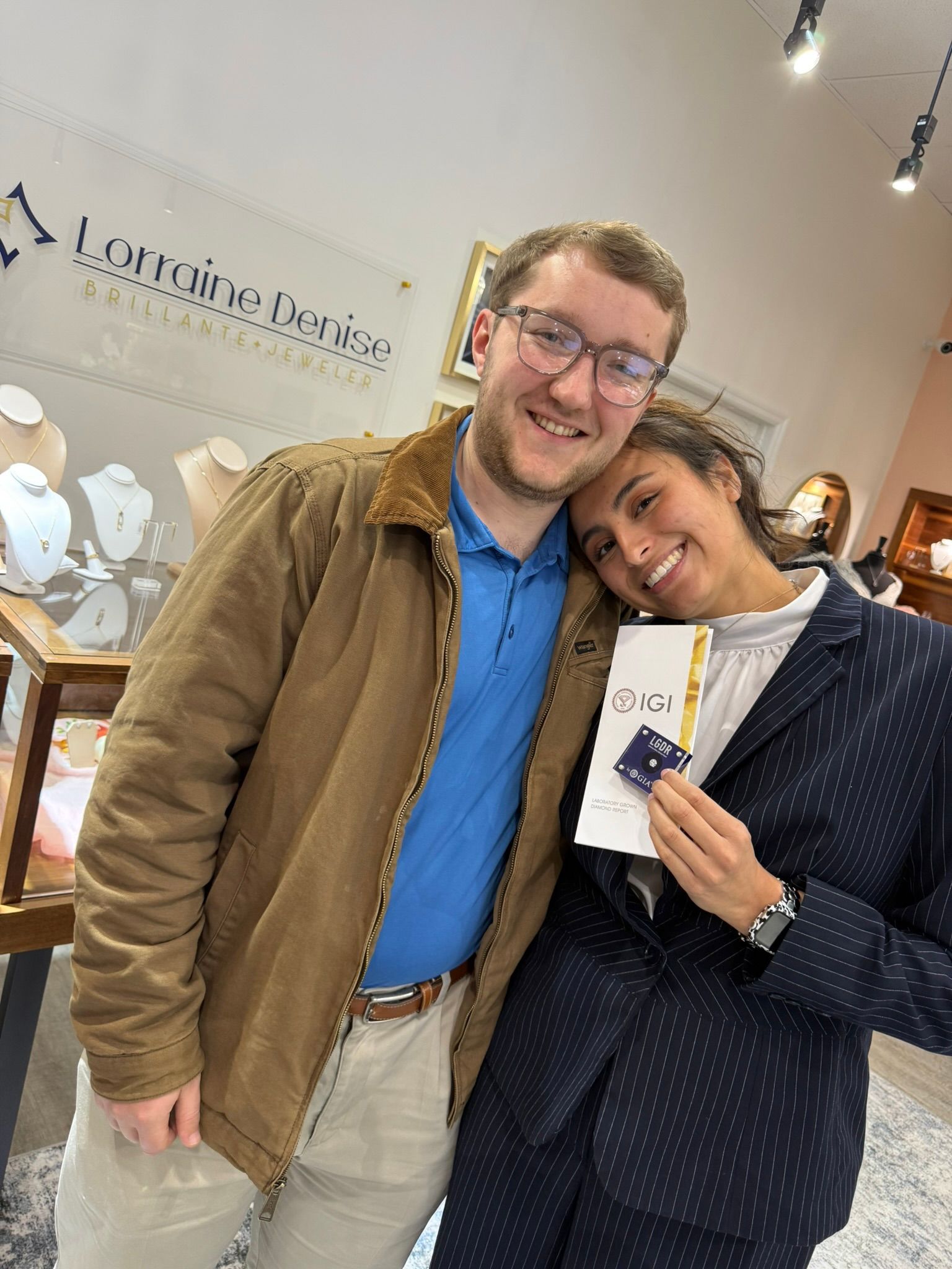 Two smiling people pose indoors, one holding a small white item, in a bright shop.