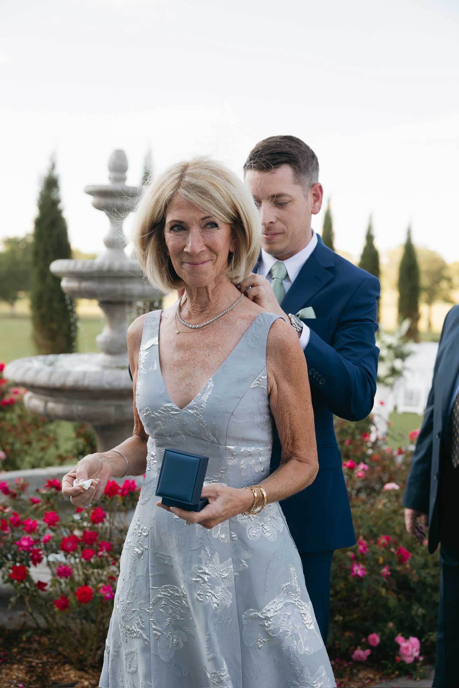 Woman in a silver dress holding a phone, with a man in a blue suit behind her by a fountain and flowers