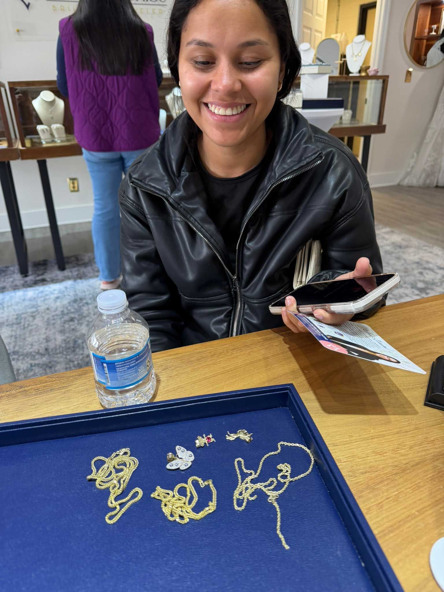 Smiling person at a table with gold jewelry and a blue tray, holding a phone