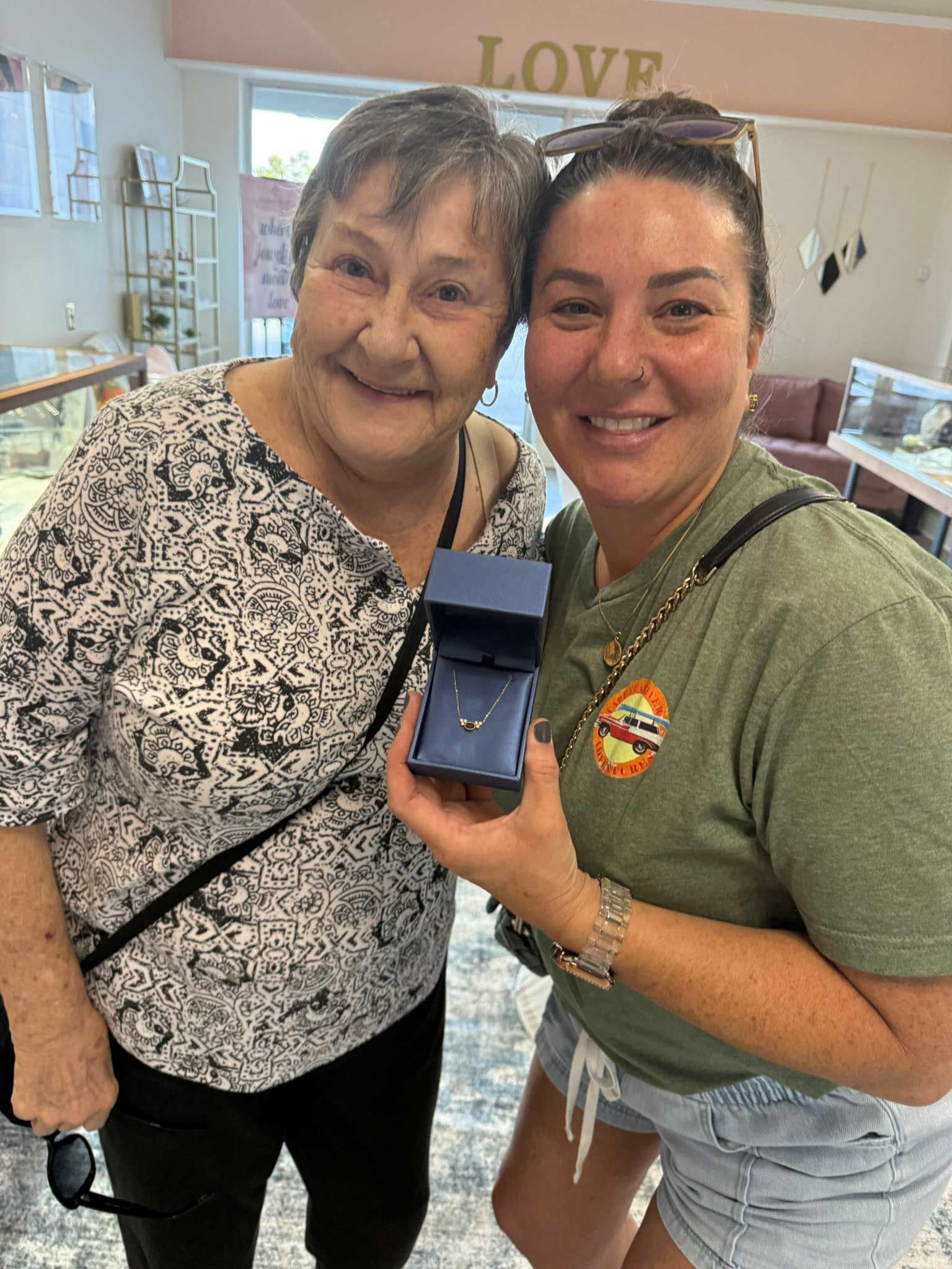 Two women smiling in a shop, holding up a blue ring box together