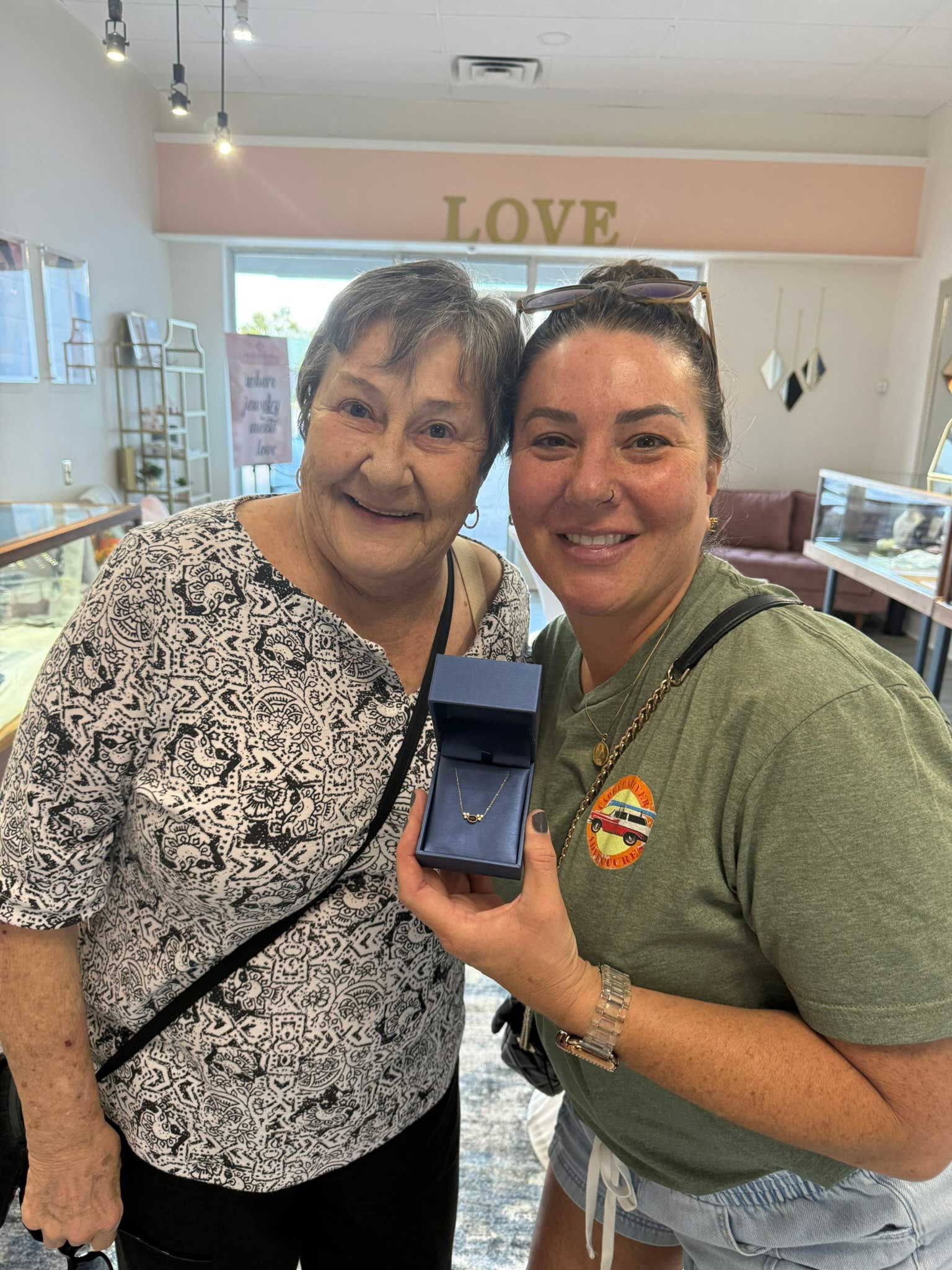 Two women smiling in a jewelry store, one holding up an open ring box.