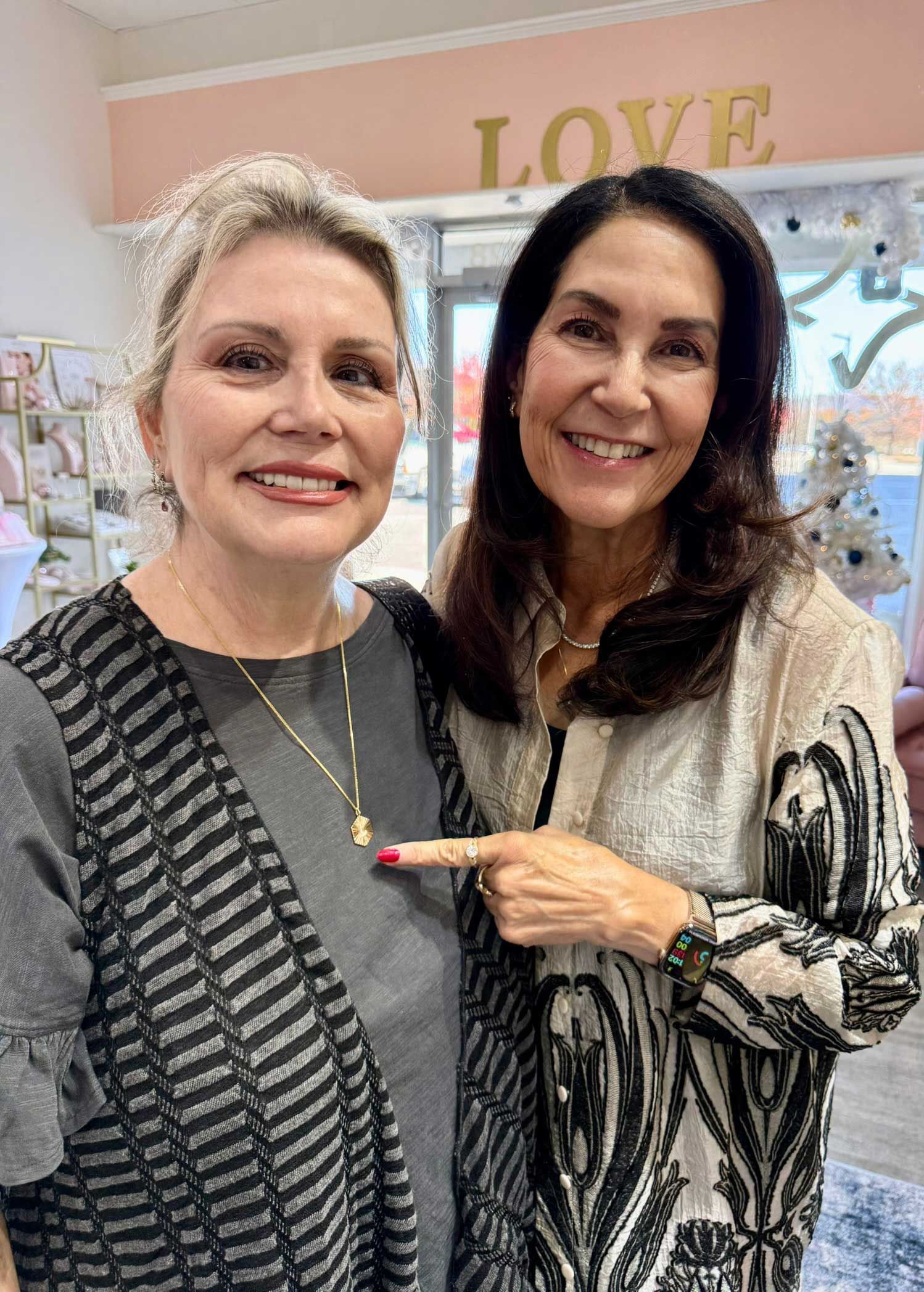 Two smiling women standing indoors, one pointing at the other in a boutique with a sign behind them