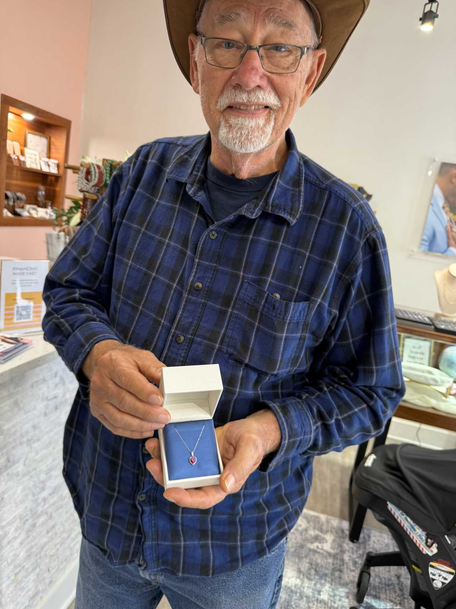 Man in a blue plaid shirt holding an open ring box with a blue stone inside in a shop.