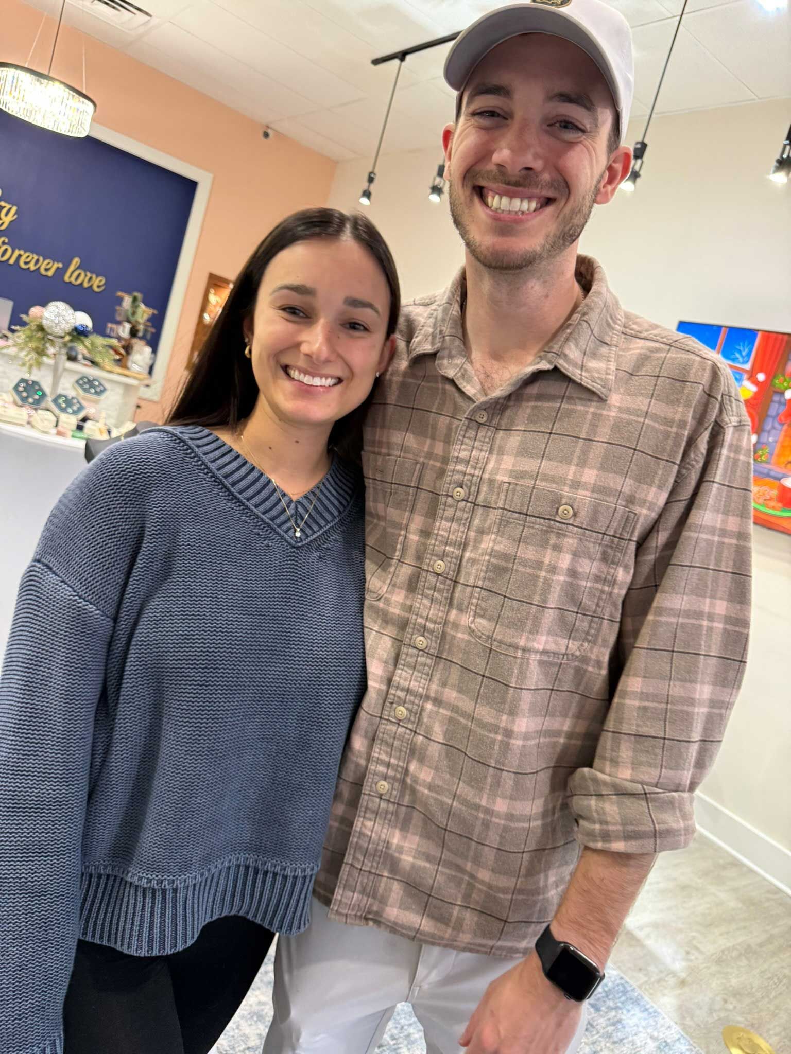 Two smiling people standing indoors, wearing casual sweaters and a cap.
