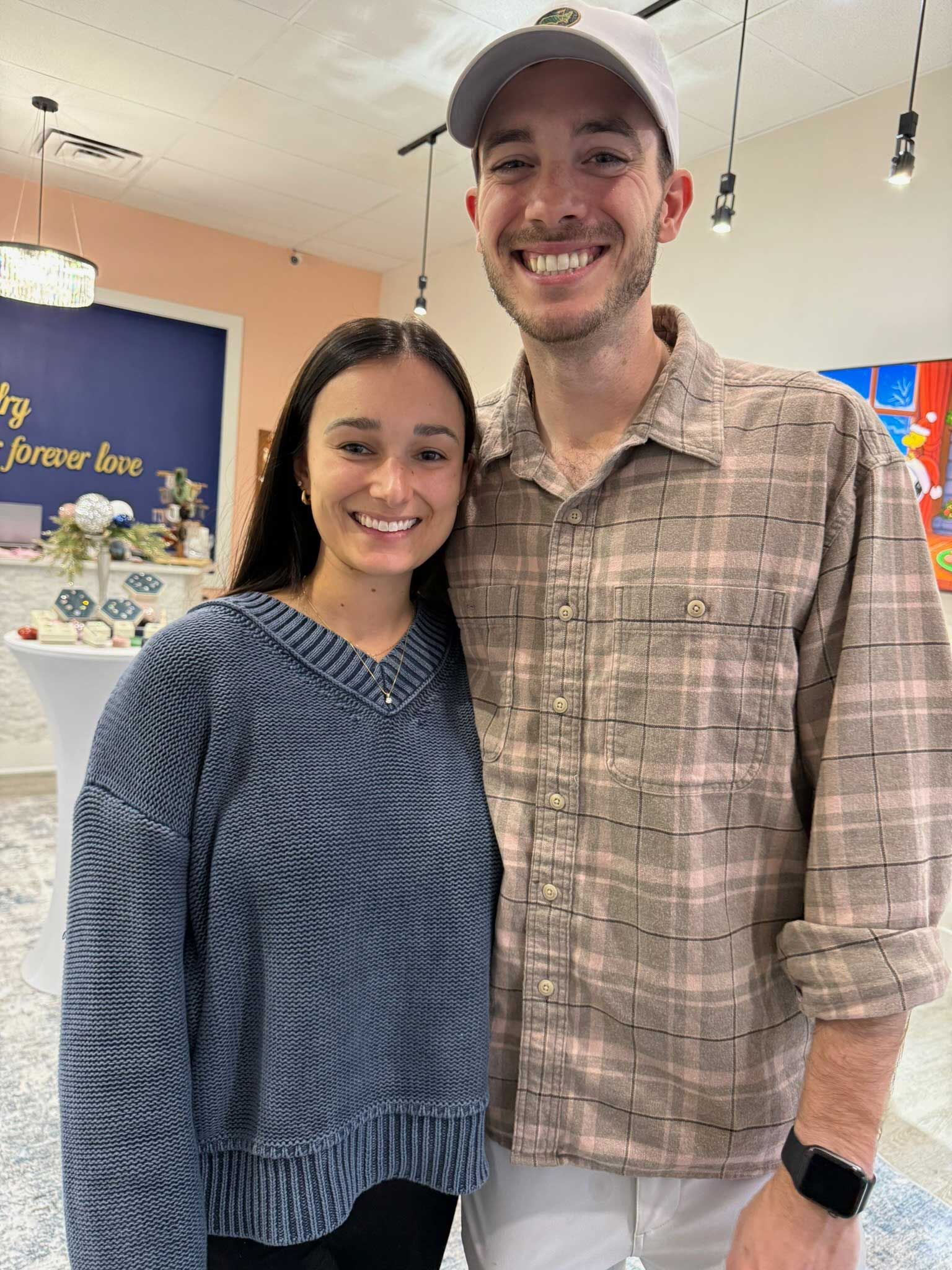Two smiling people standing indoors, wearing casual plaid and blue patterned tops.