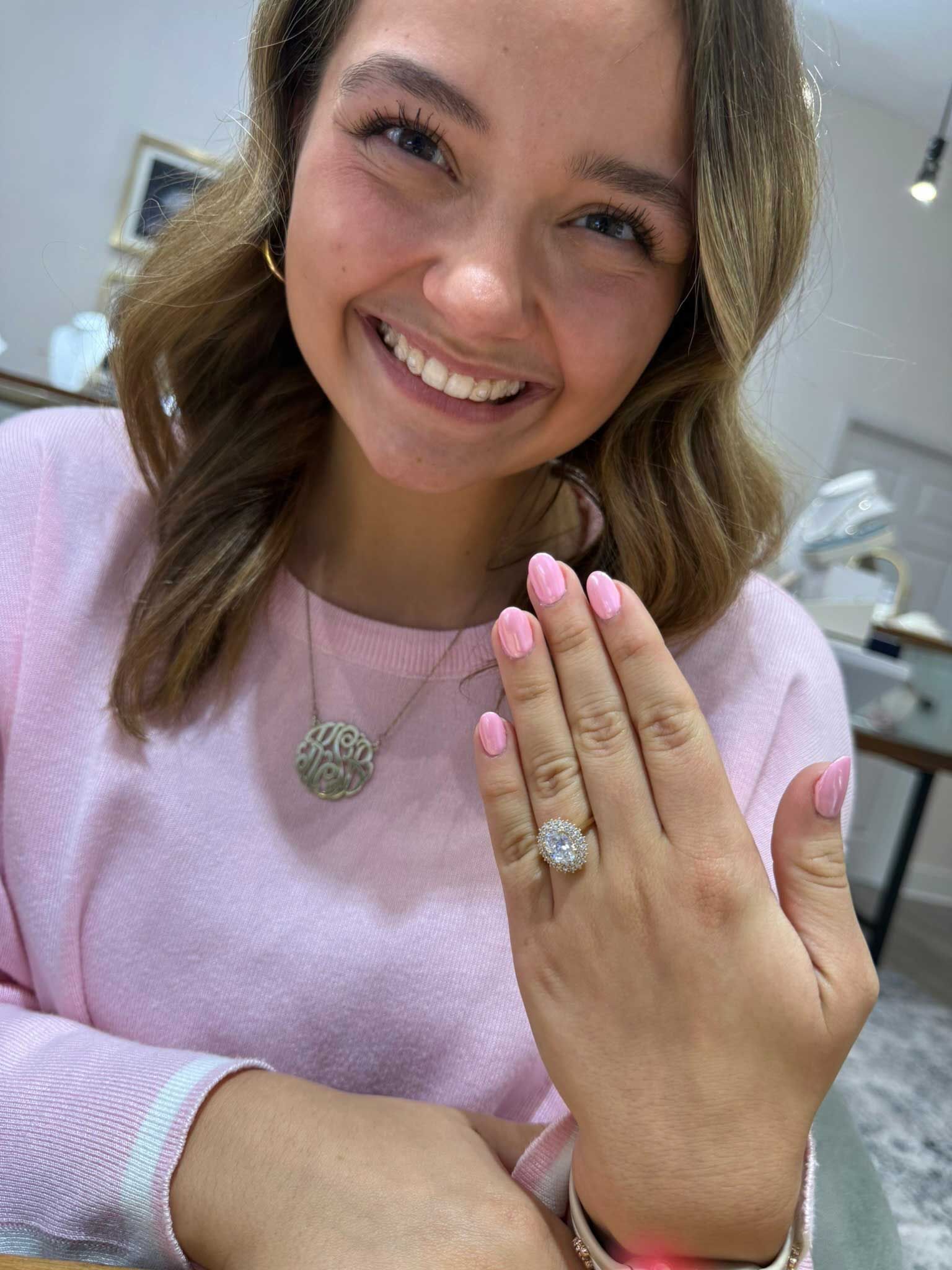 Smiling woman showing a hand with a large ring and pink nails, wearing a light pink sweater.