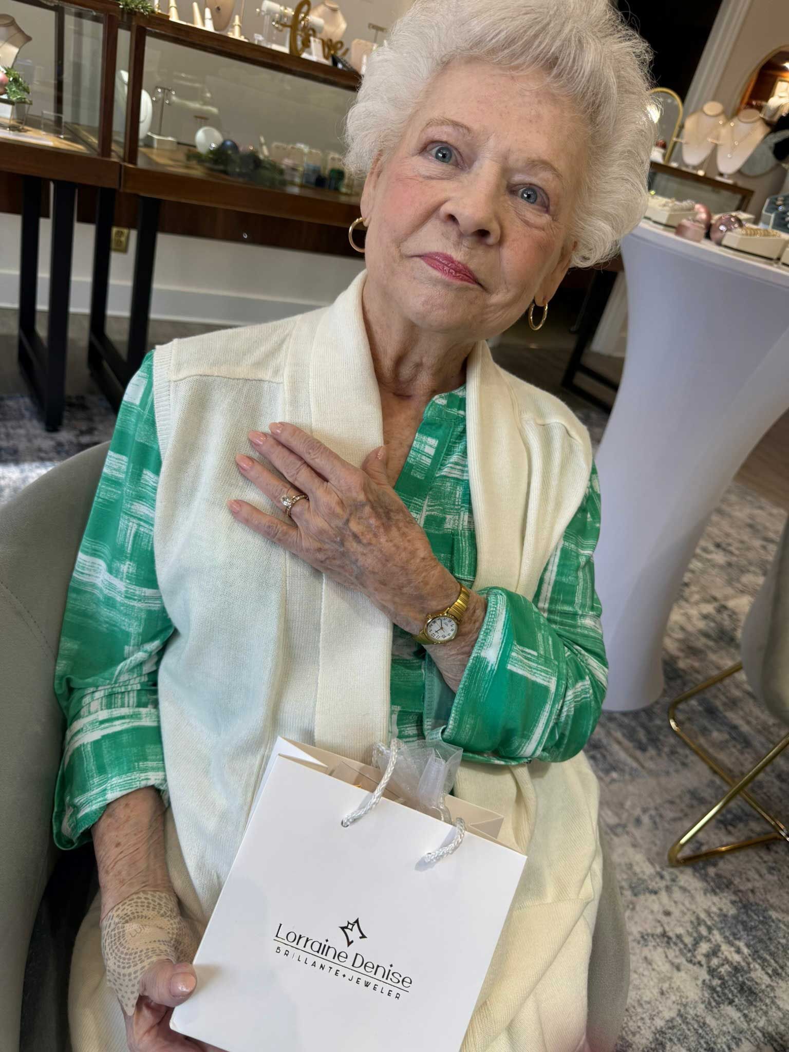 Elderly woman seated indoors, wearing a green patterned outfit and cream shawl, holding a white gift bag.