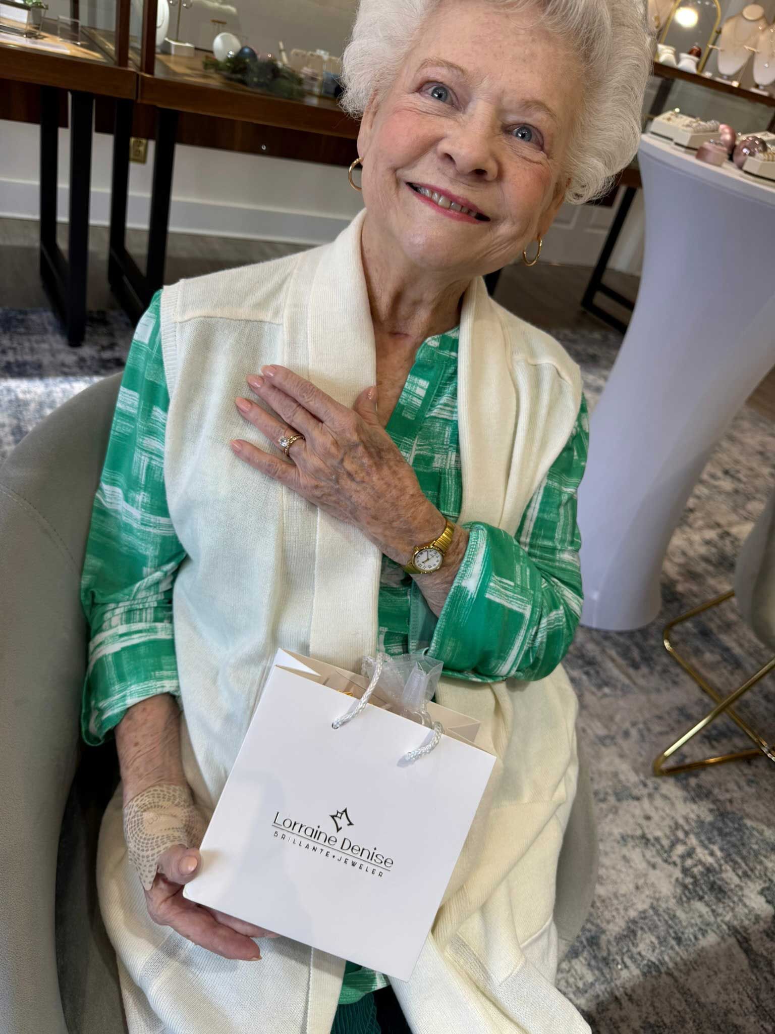 Elderly woman in a green patterned outfit smiling and holding a white gift bag in a formal indoor setting