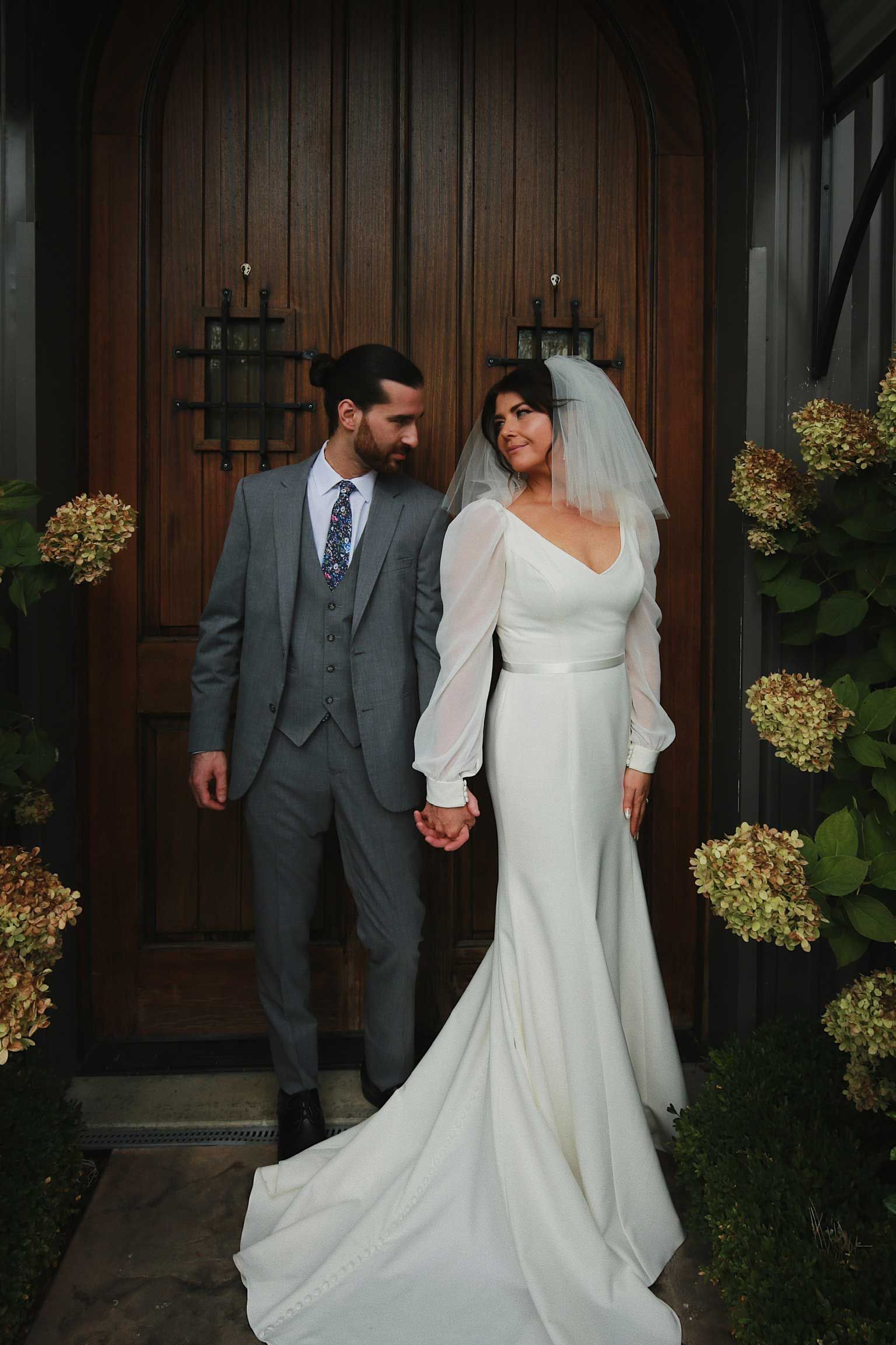 Bride and groom holding hands in formal attire outside wooden doors, bride in white gown and veil
