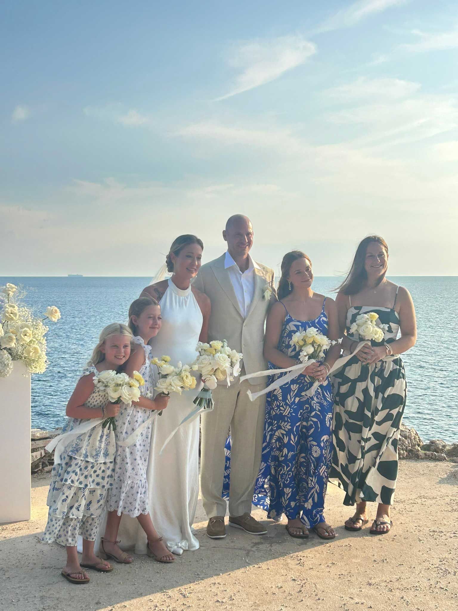 Five people posing with bouquets by the sea at sunset, wearing formal and floral outfits.