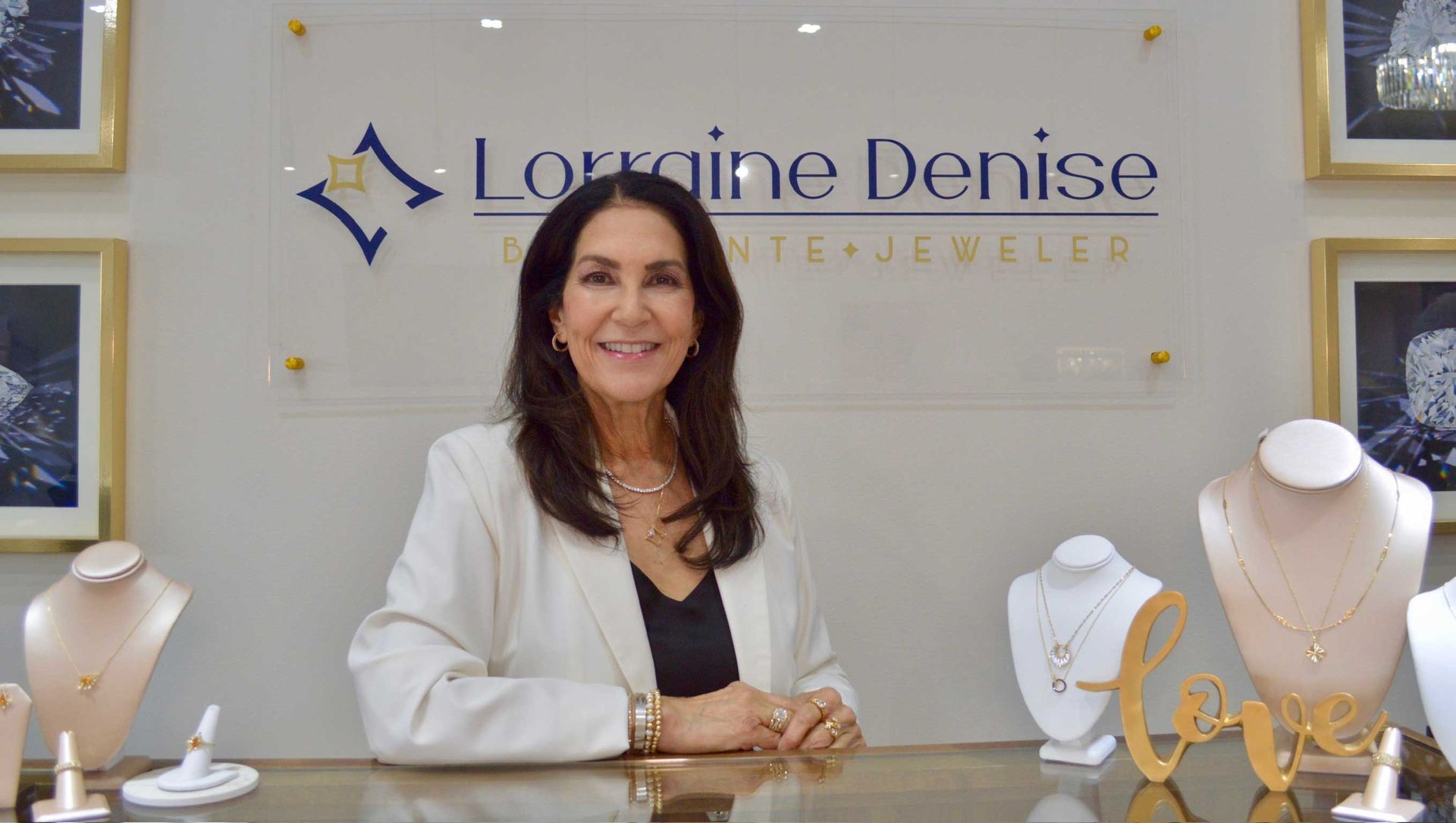 Jewelry boutique owner standing behind display cases with necklaces and rings, in front of a sign