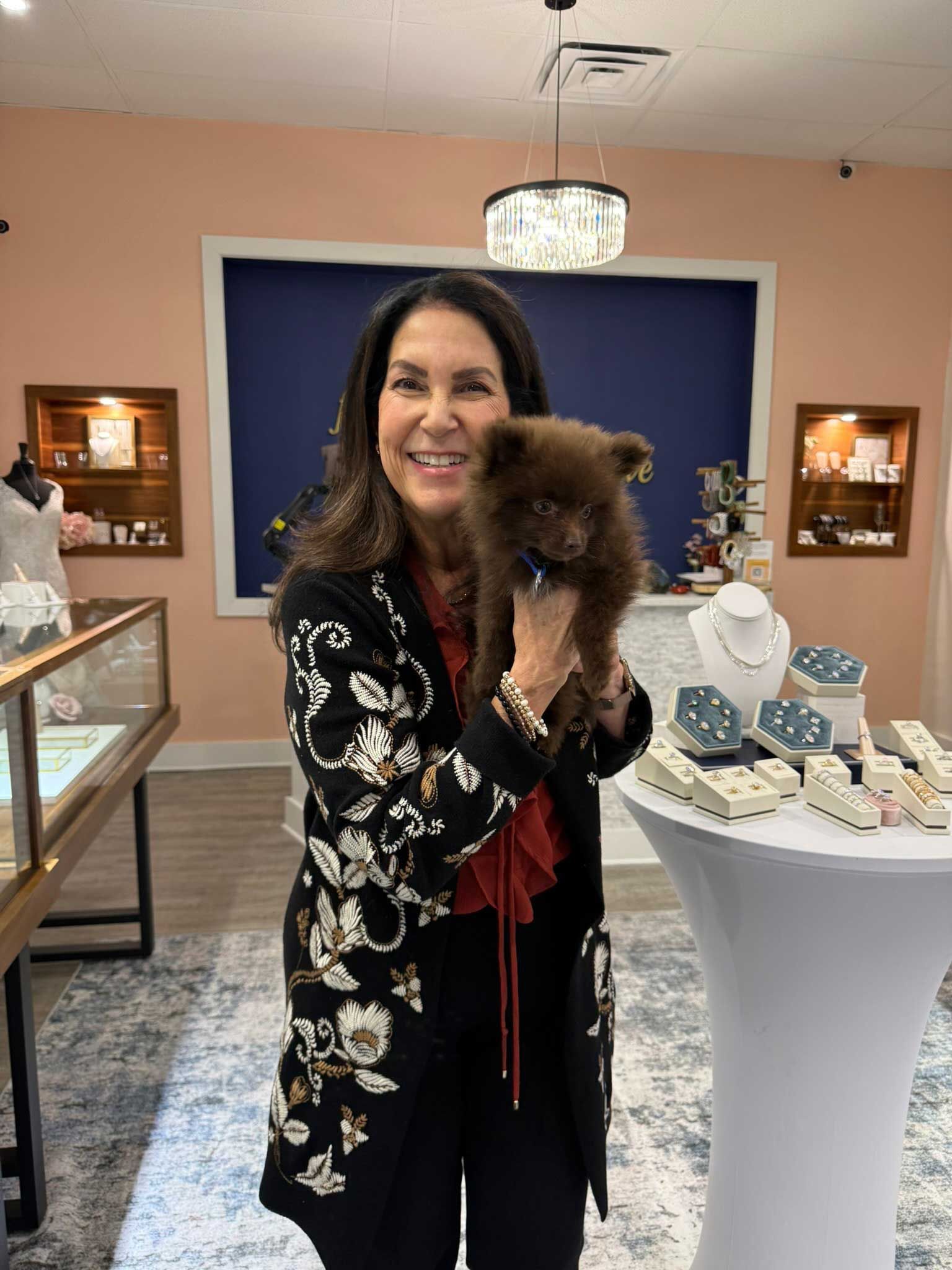 Woman holding a small brown dog at an indoor boutique display, smiling at the camera.