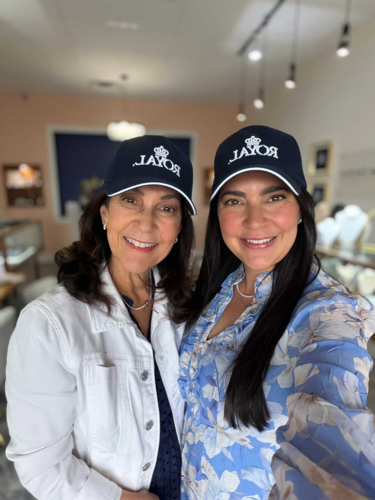 Two women smiling in black caps, posing indoors, one in a white jacket and one in a blue floral shirt.