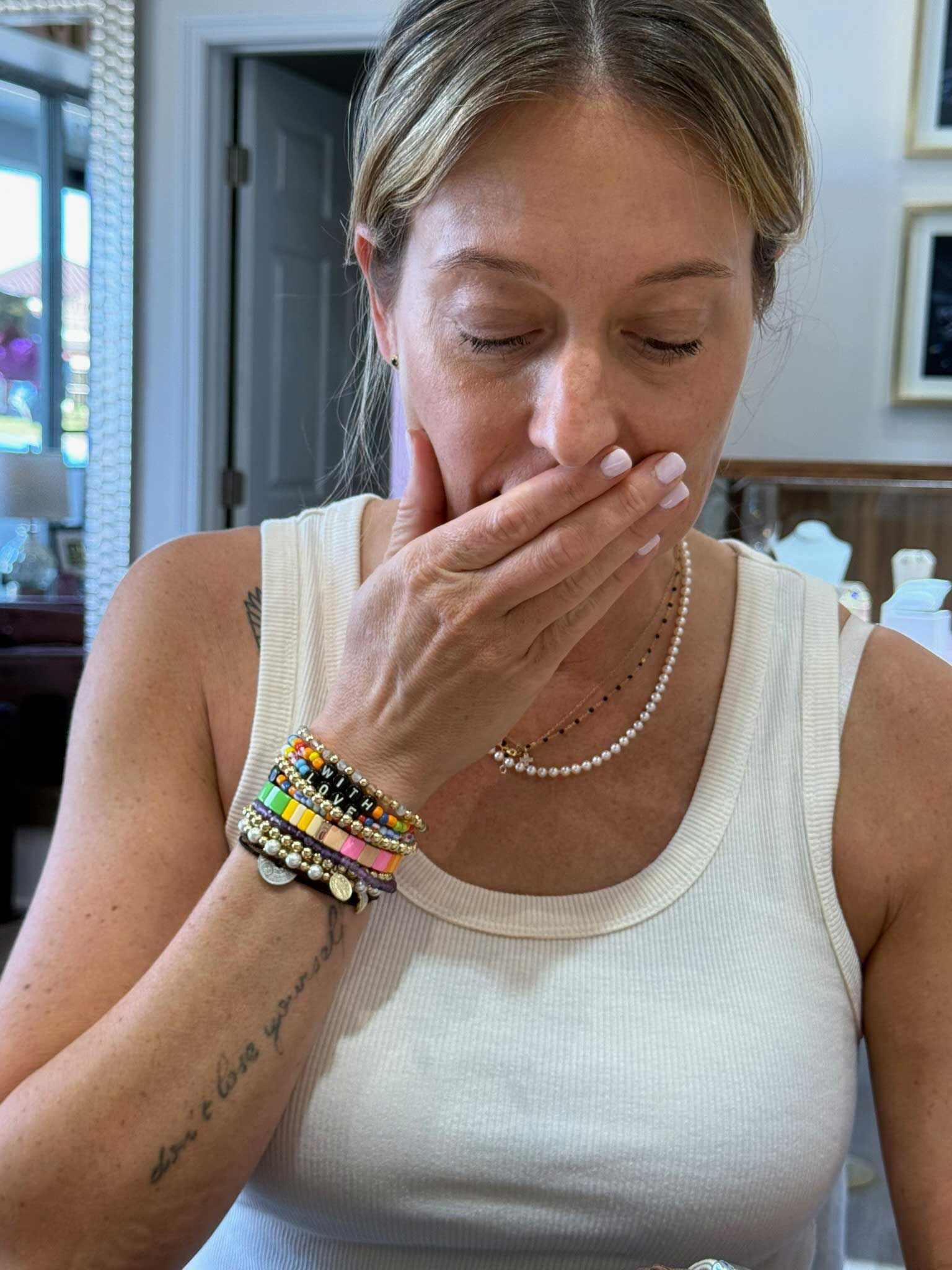 Woman in white tank top covering her mouth with her hand indoors, wearing colorful bracelets and a necklace