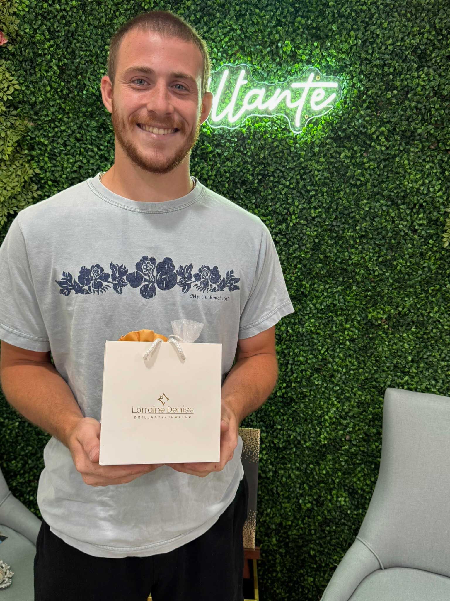 Smiling man holding a white gift bag in front of a green plant wall