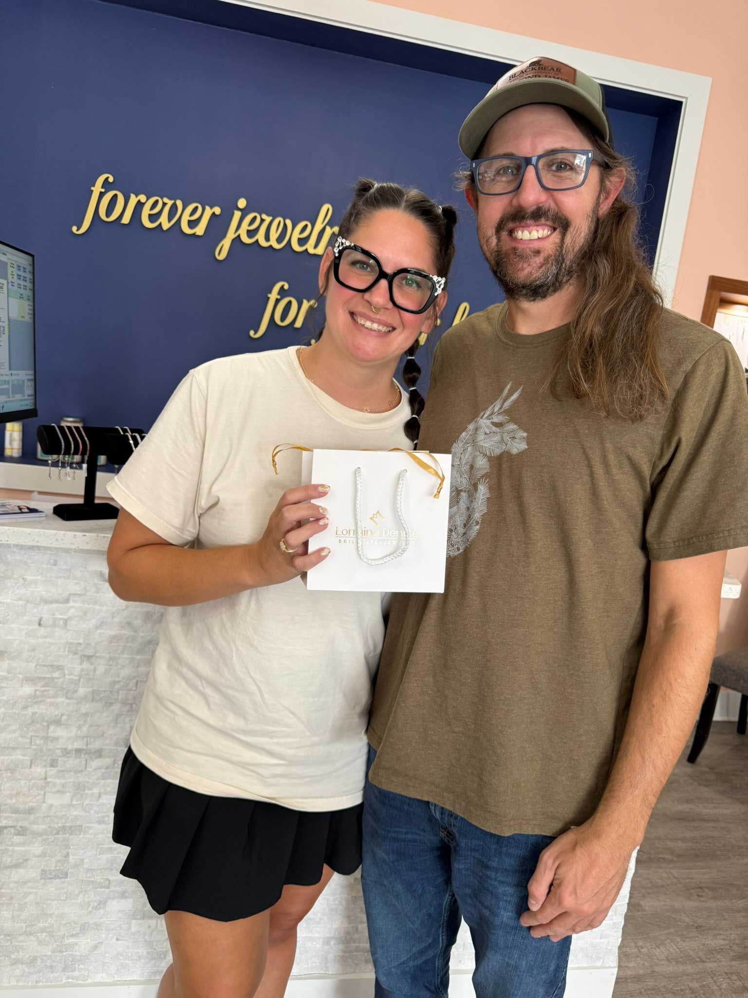 Two smiling people in a jewelry shop, holding a necklace display card with a blue wall sign behind them.