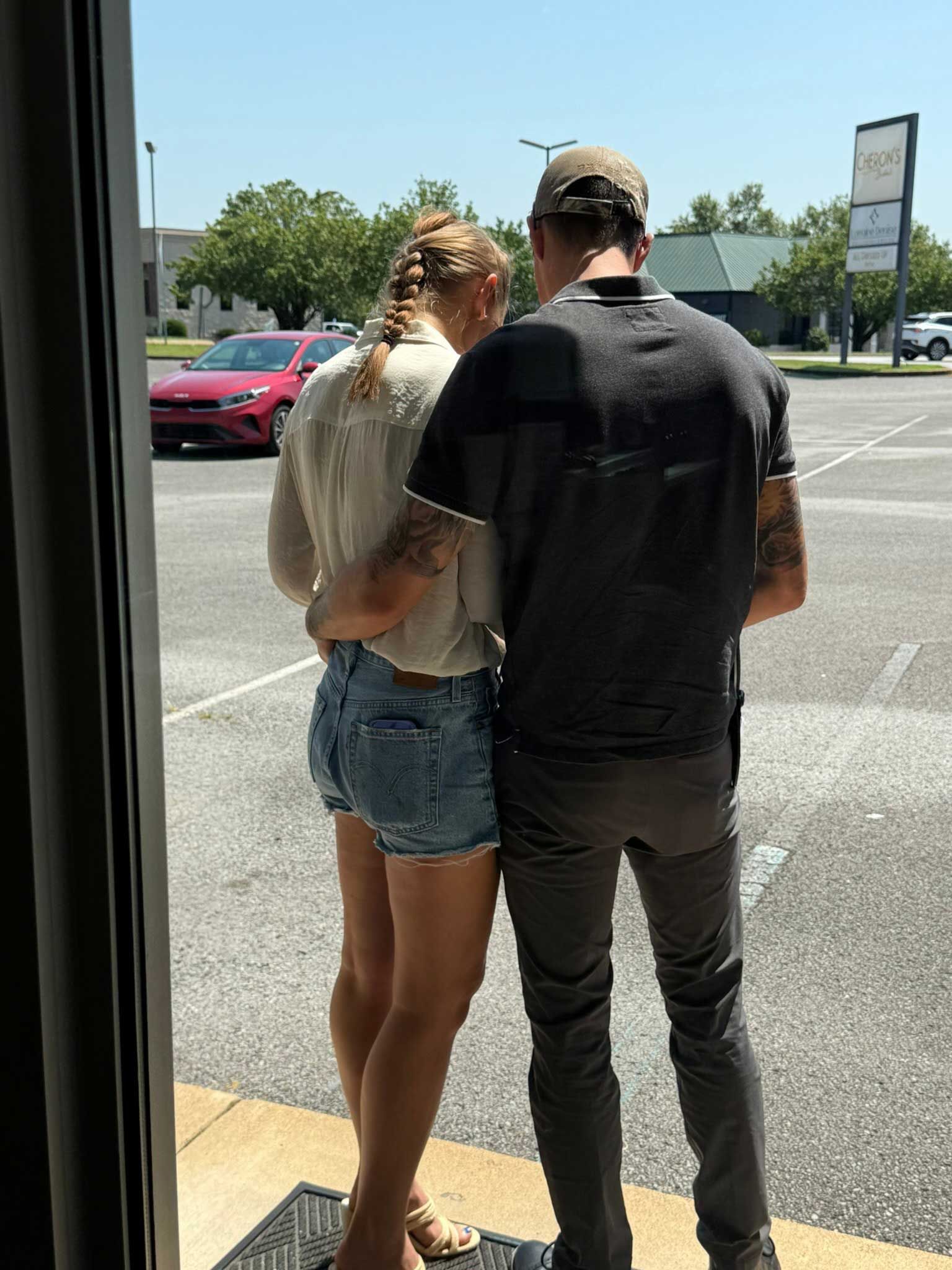 Two people standing close together in a sunny parking lot, seen from behind.