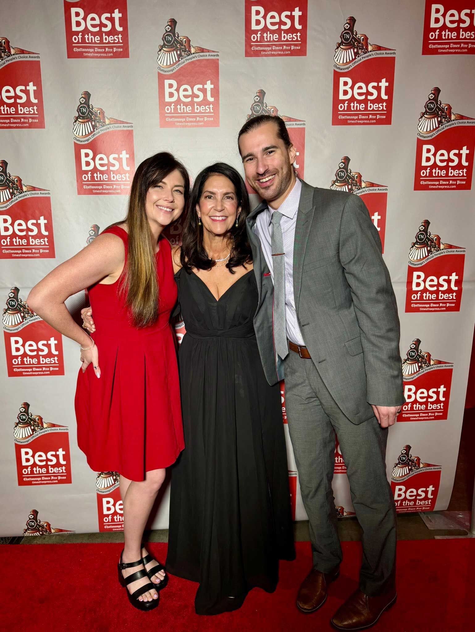 Three people posing on a red carpet in front of a Best Awards backdrop, smiling and dressed formally