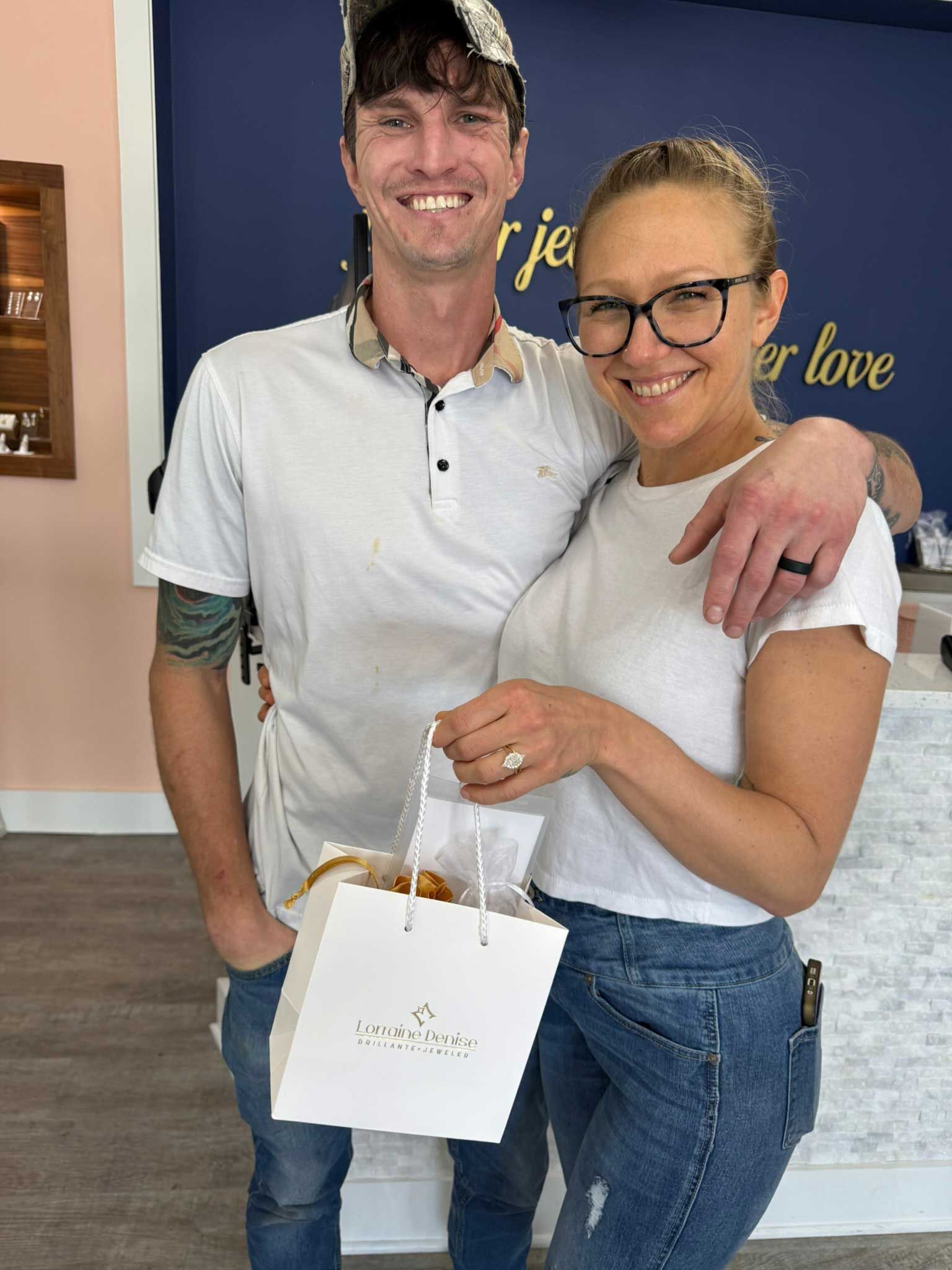 Smiling couple holding a white gift bag in front of a wall sign.