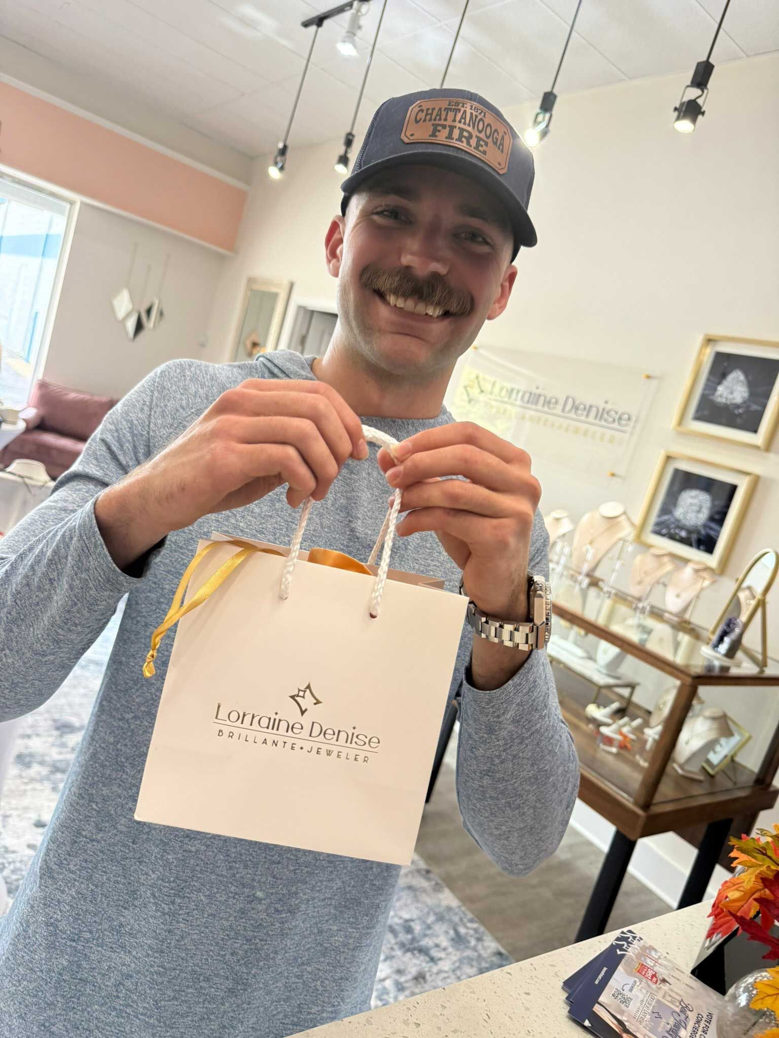Smiling man holding a white gift bag in a jewelry store, with display cases and framed art behind him.
