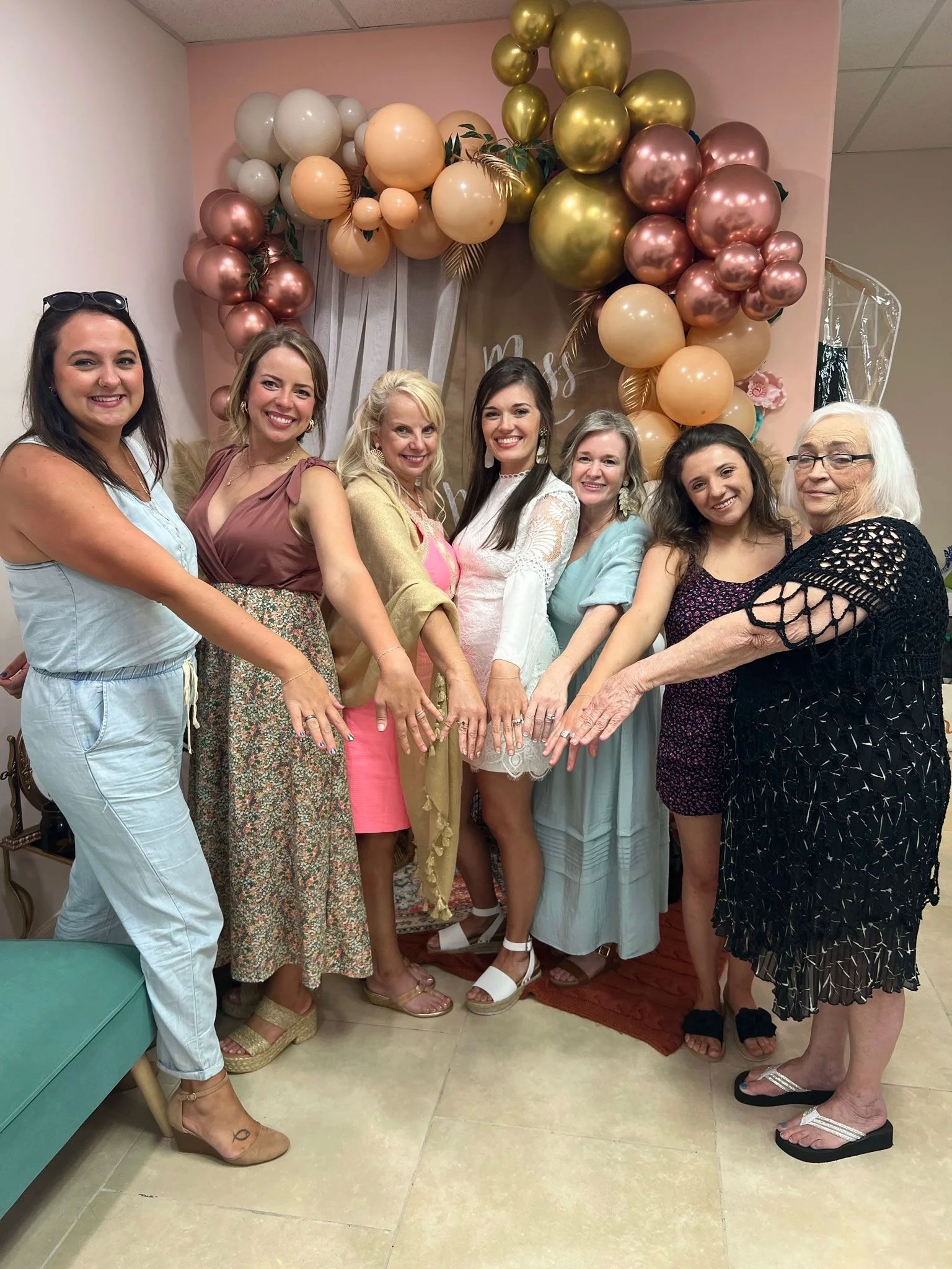 Seven women with hands together in front of a balloon arch; celebratory event.