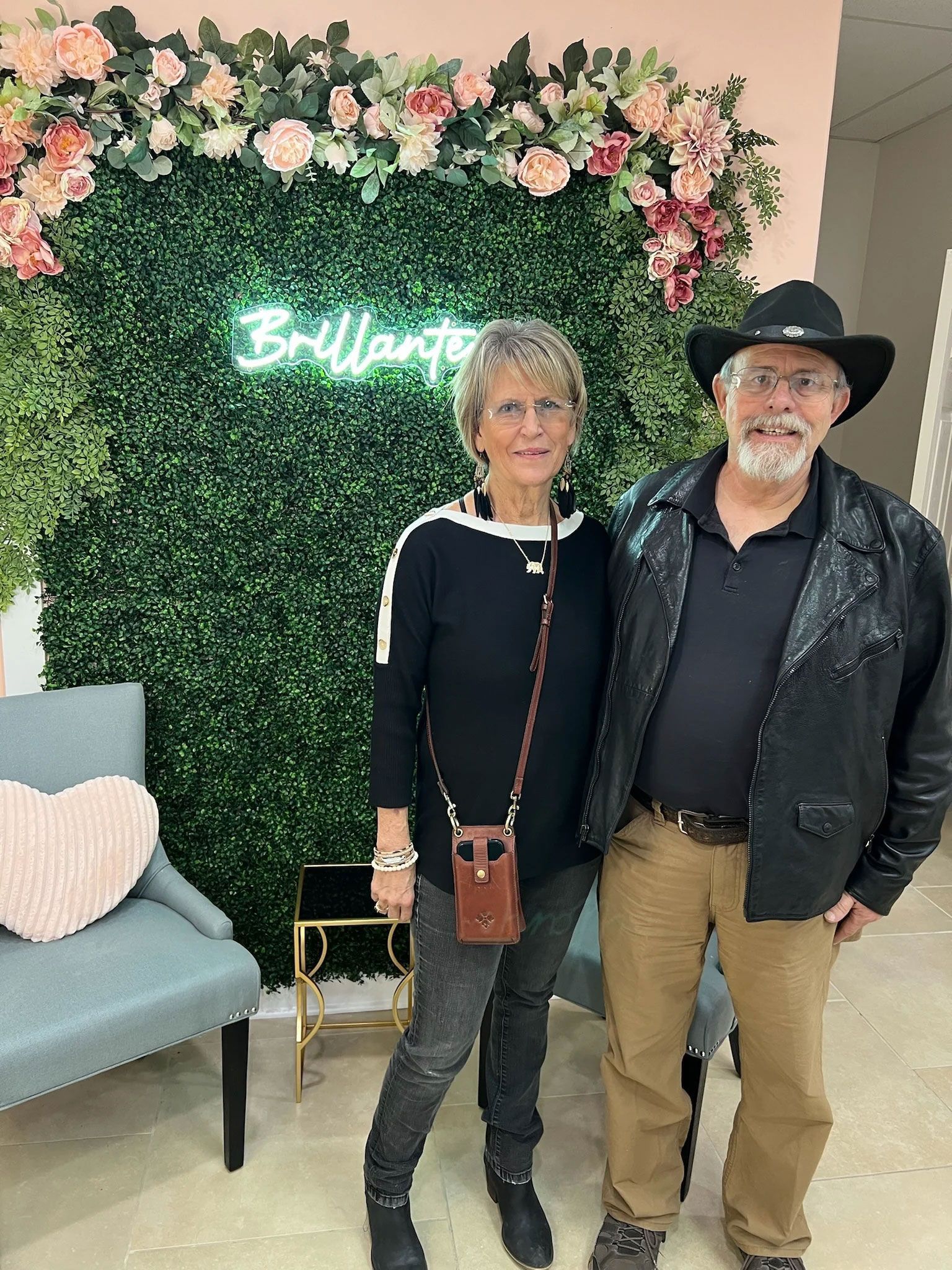 Couple poses in front of a green wall with flowers and neon sign, woman in black top and jeans, man in cowboy hat.