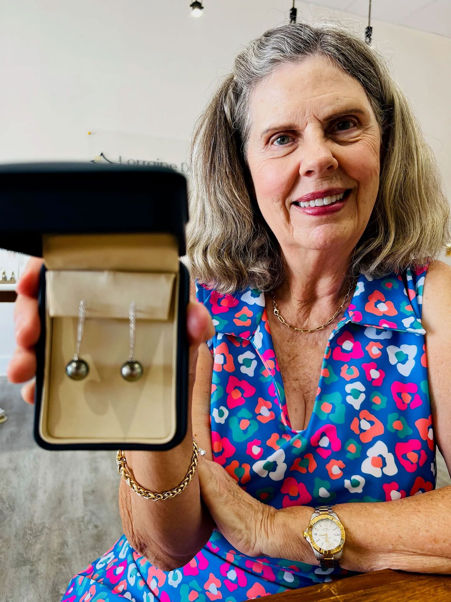 Woman holding a box with black pearl earrings, smiling, wearing a colorful dress.