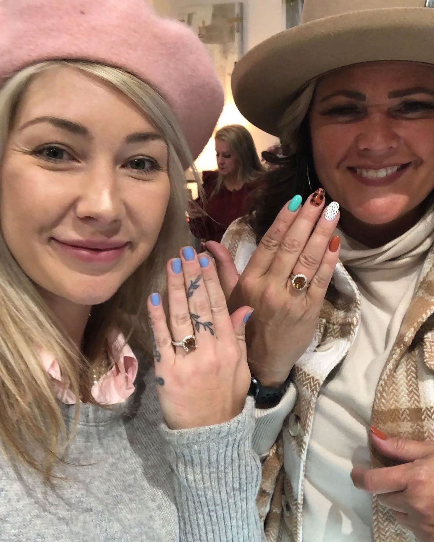 Two smiling women showing off rings, one with a pink beret, the other a hat, both with manicured nails.