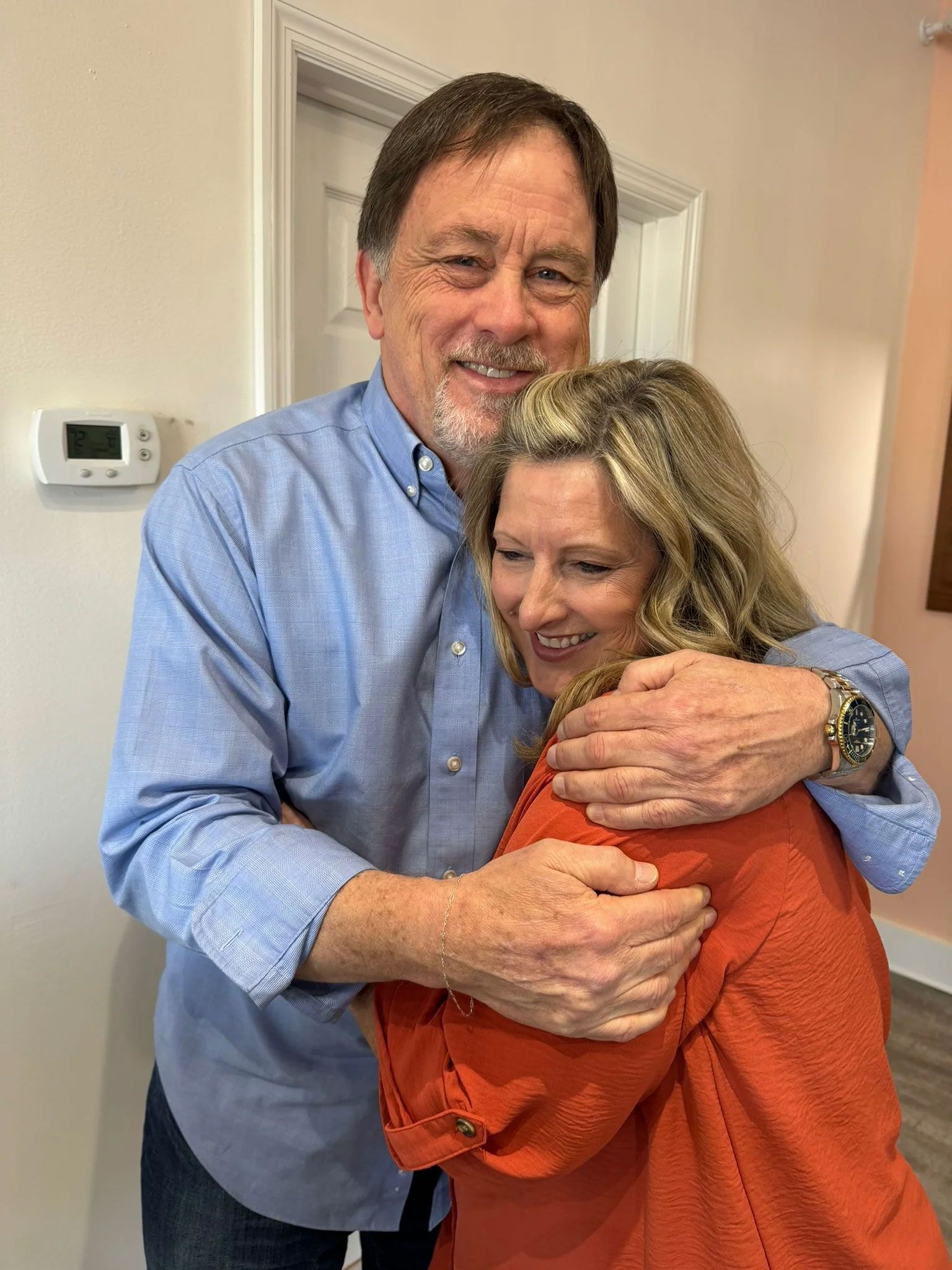 Man in blue shirt embraces a woman in orange. They smile indoors, near a door.